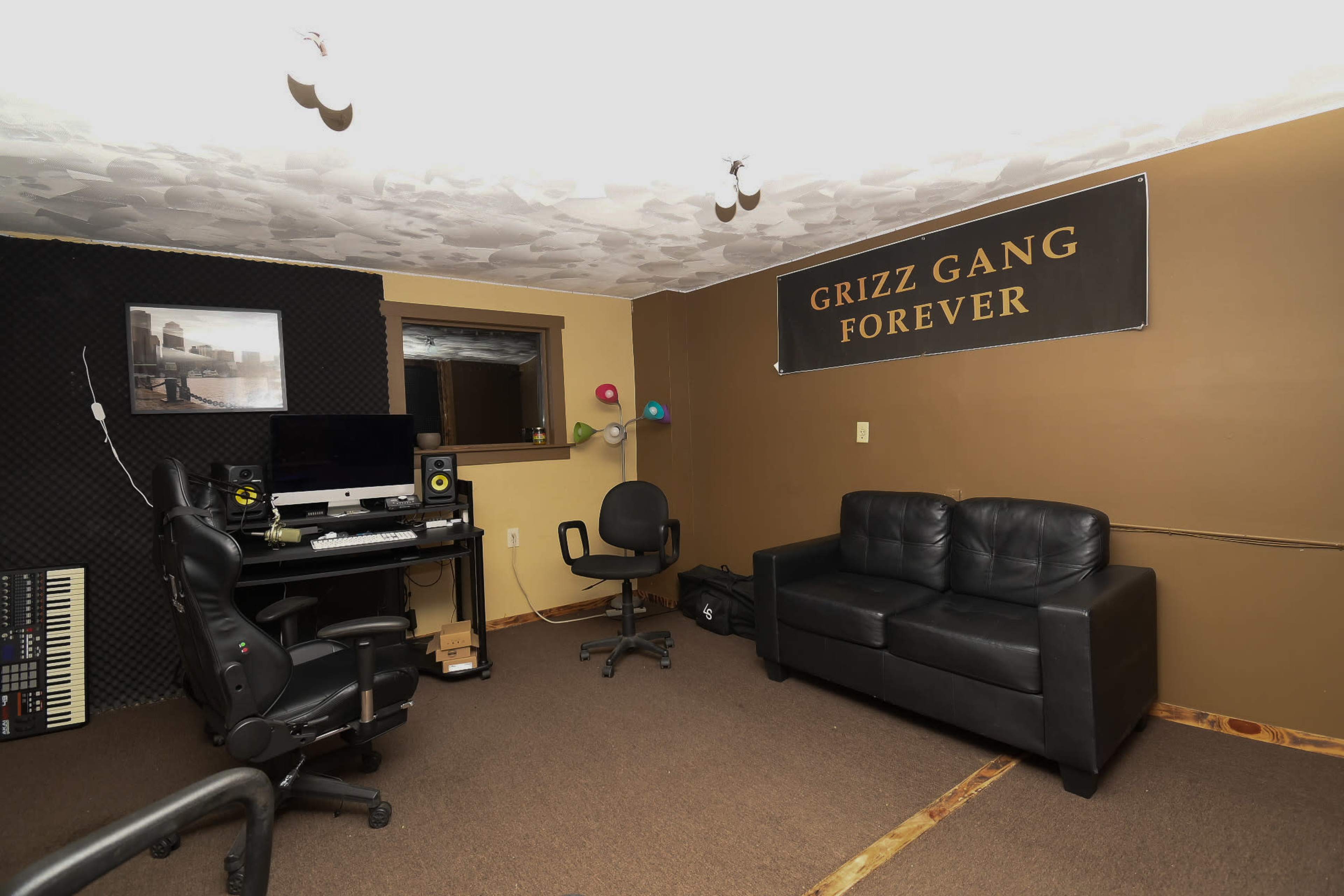 The image shows a small room with a black leather couch, two office chairs, a computer setup with speakers, and a banner reading "GRIZZ GANG FOREVER" on the wall.