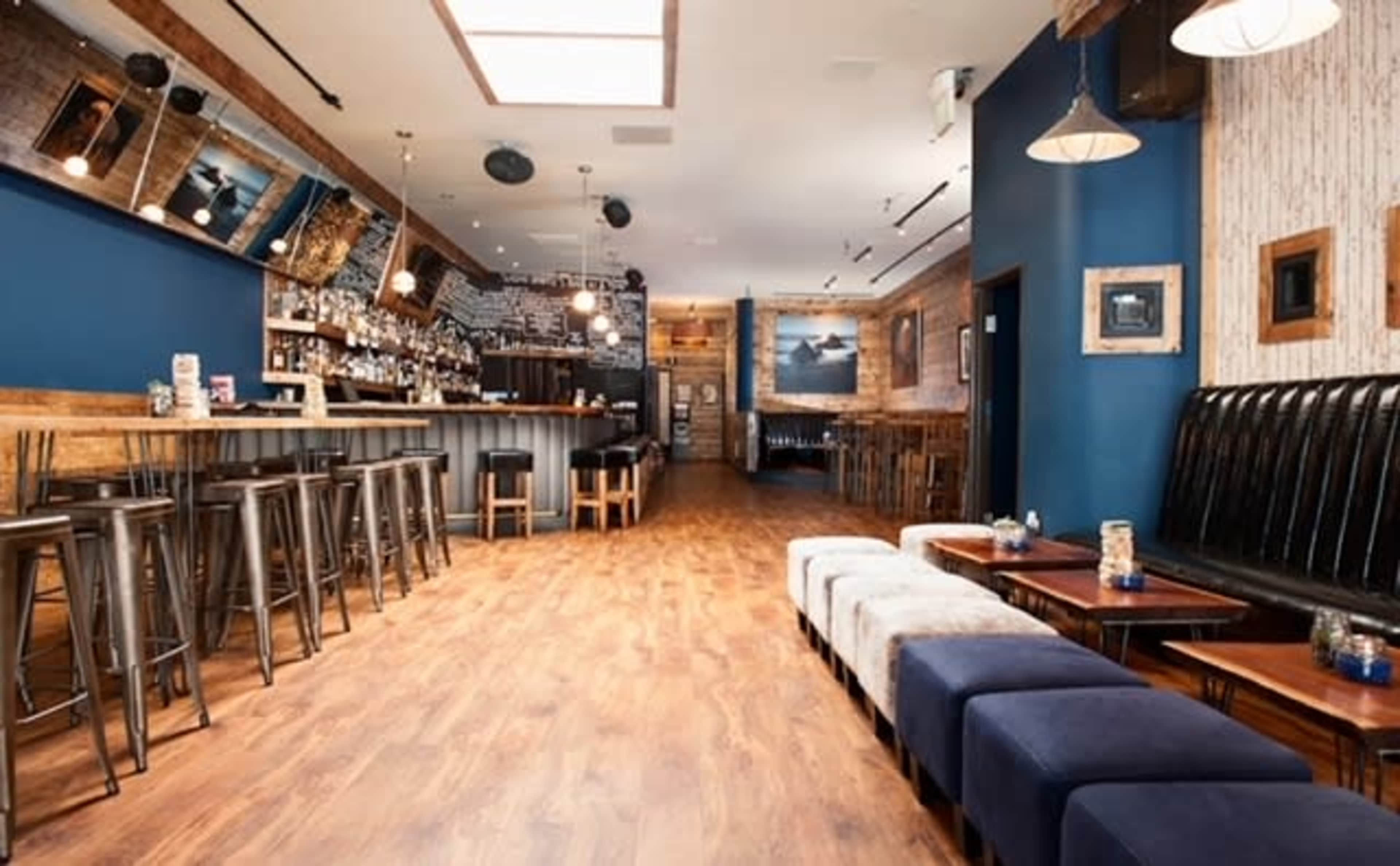 A modern bar interior with a wooden floor, a long bar counter, and multiple seating areas featuring a mix of stools and benches.