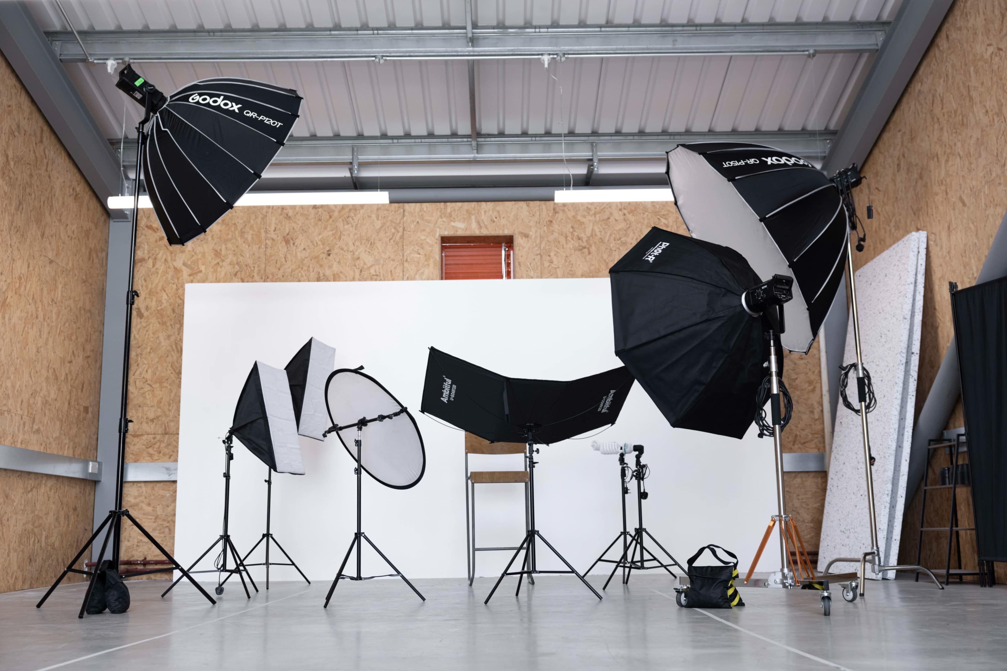 The image shows a photography studio equipped with various lighting setups, including softboxes and reflectors, arranged around a blank backdrop.
