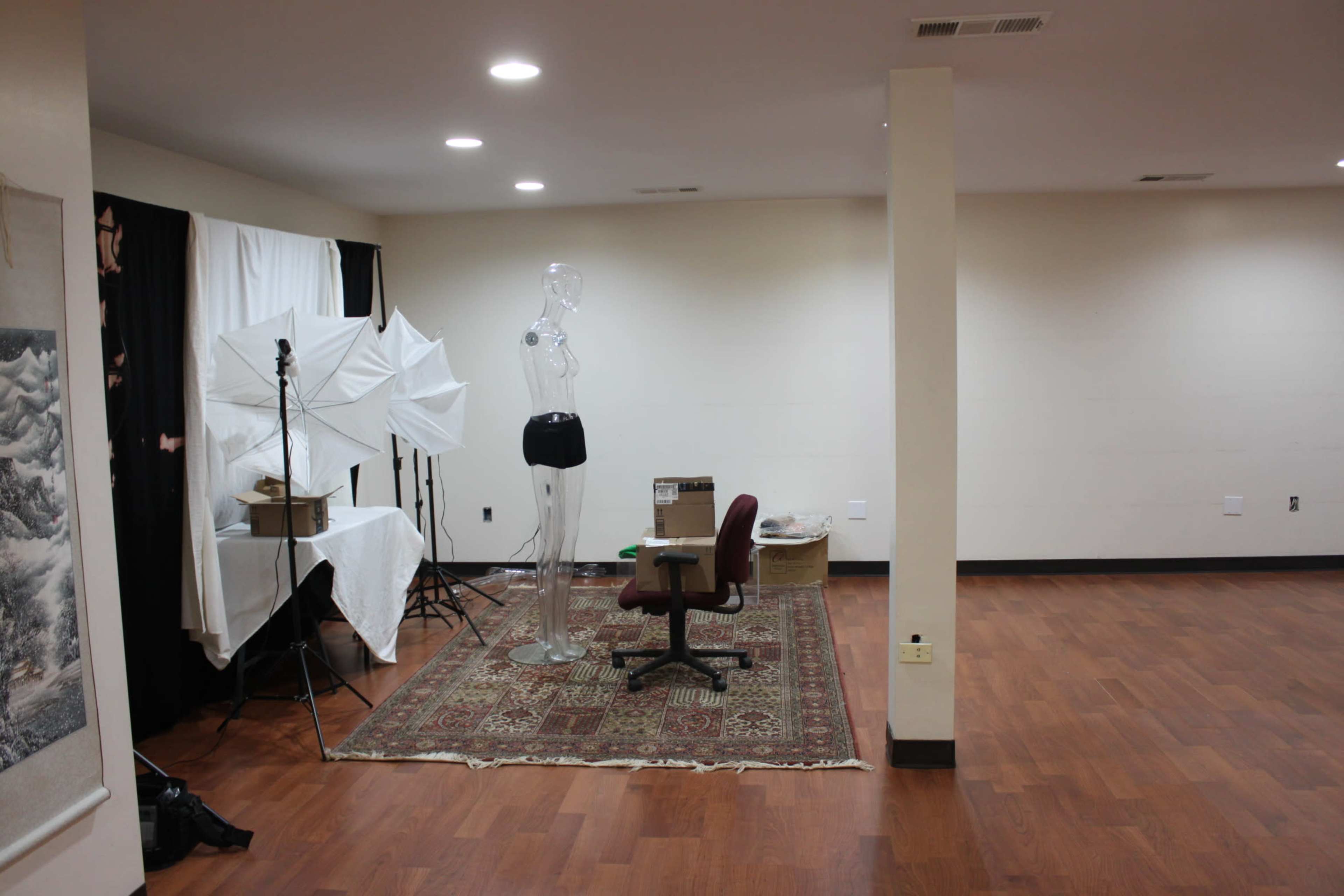 The image shows an empty room featuring a clear mannequin, a rolling chair, and photography equipment set up against one wall, with a patterned rug on the floor.