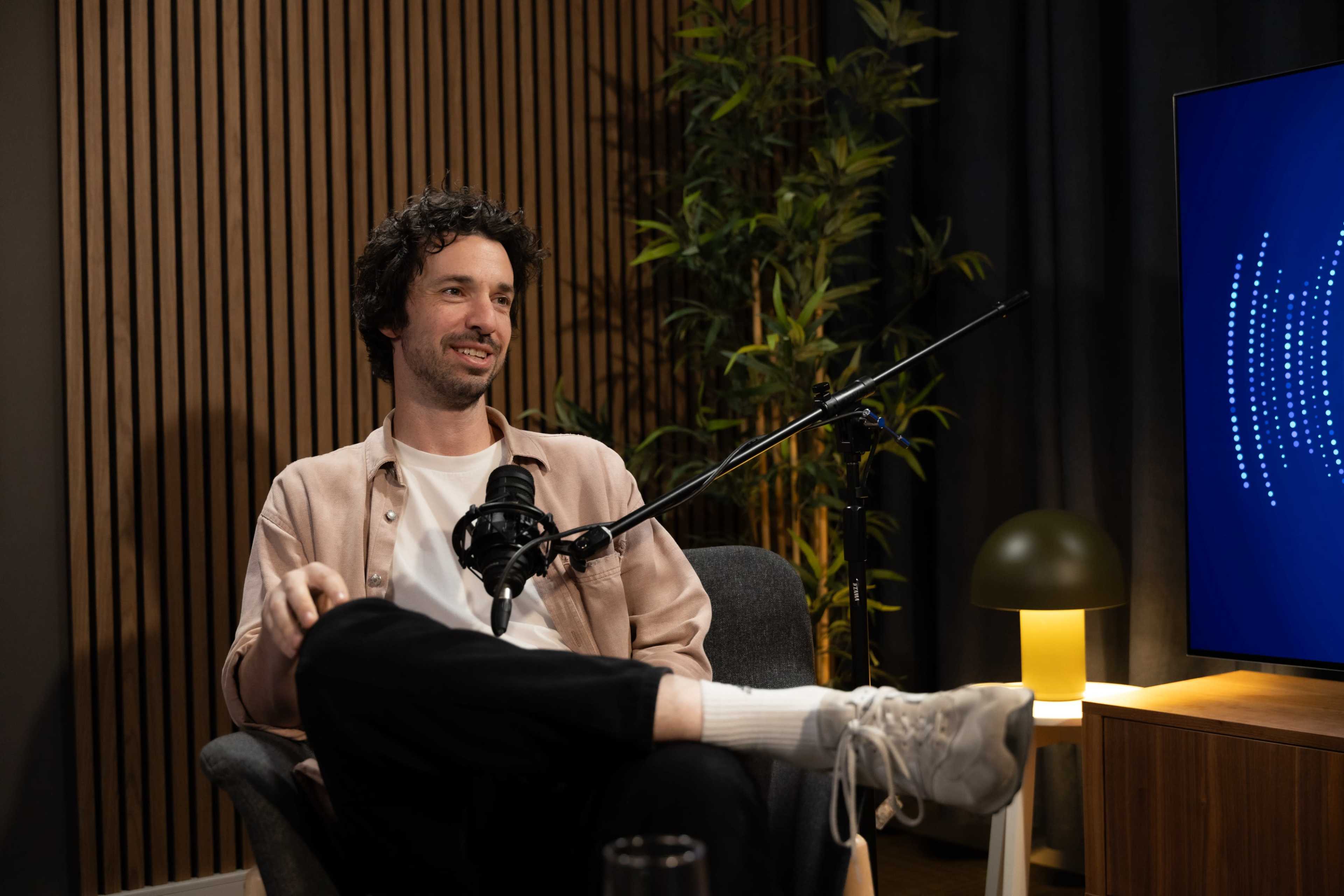 A man with curly hair sits in a chair, smiling and resting one leg over the other, in front of a microphone and a screen displaying sound waves.