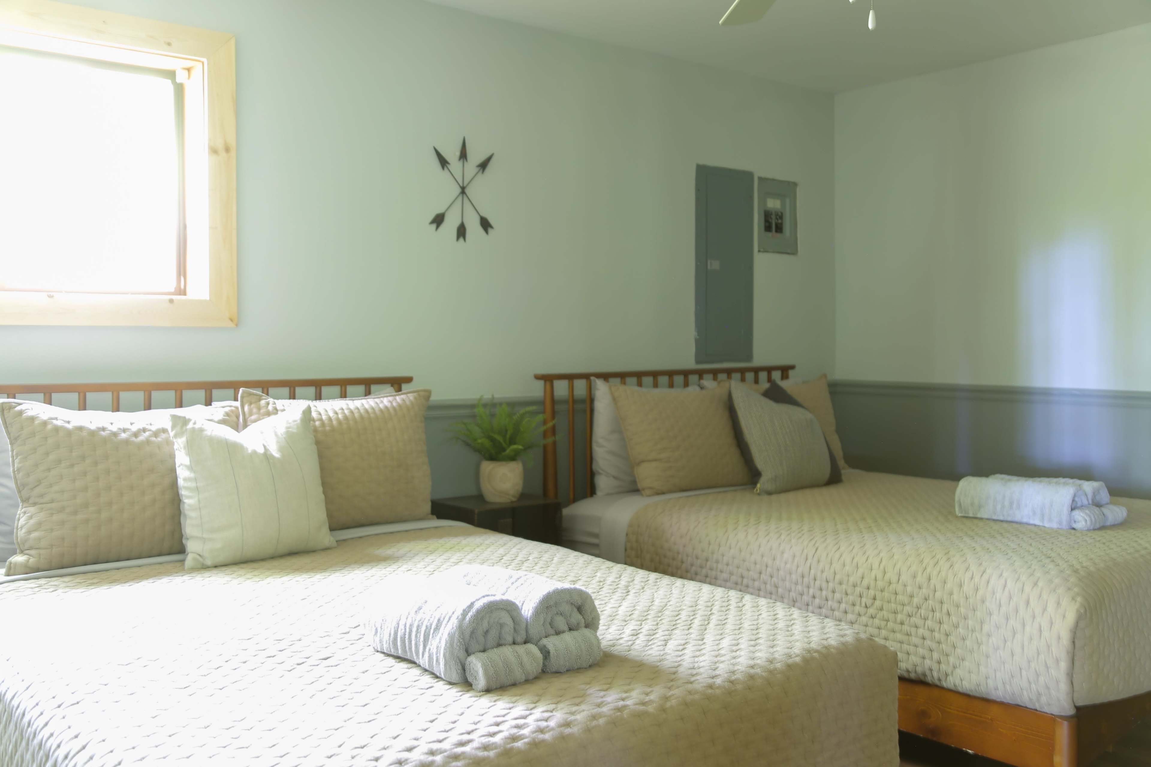 A well-lit bedroom features two double beds with neutral linens, a small potted plant, and a wall-mounted clock.