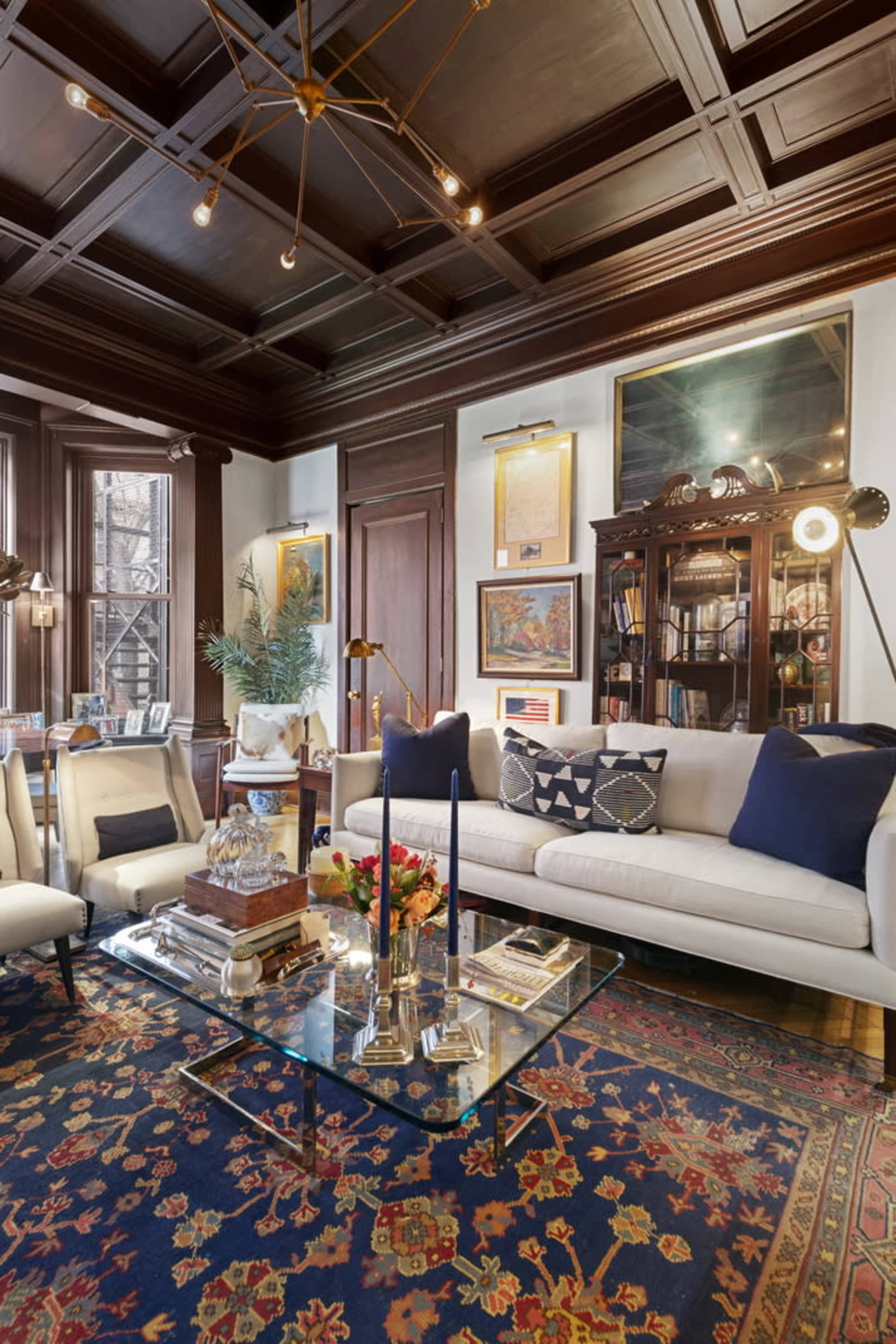 A well-furnished living room with a coffered ceiling, a beige sofa, a glass coffee table, and a vintage-style shelving unit filled with books and decorative items.