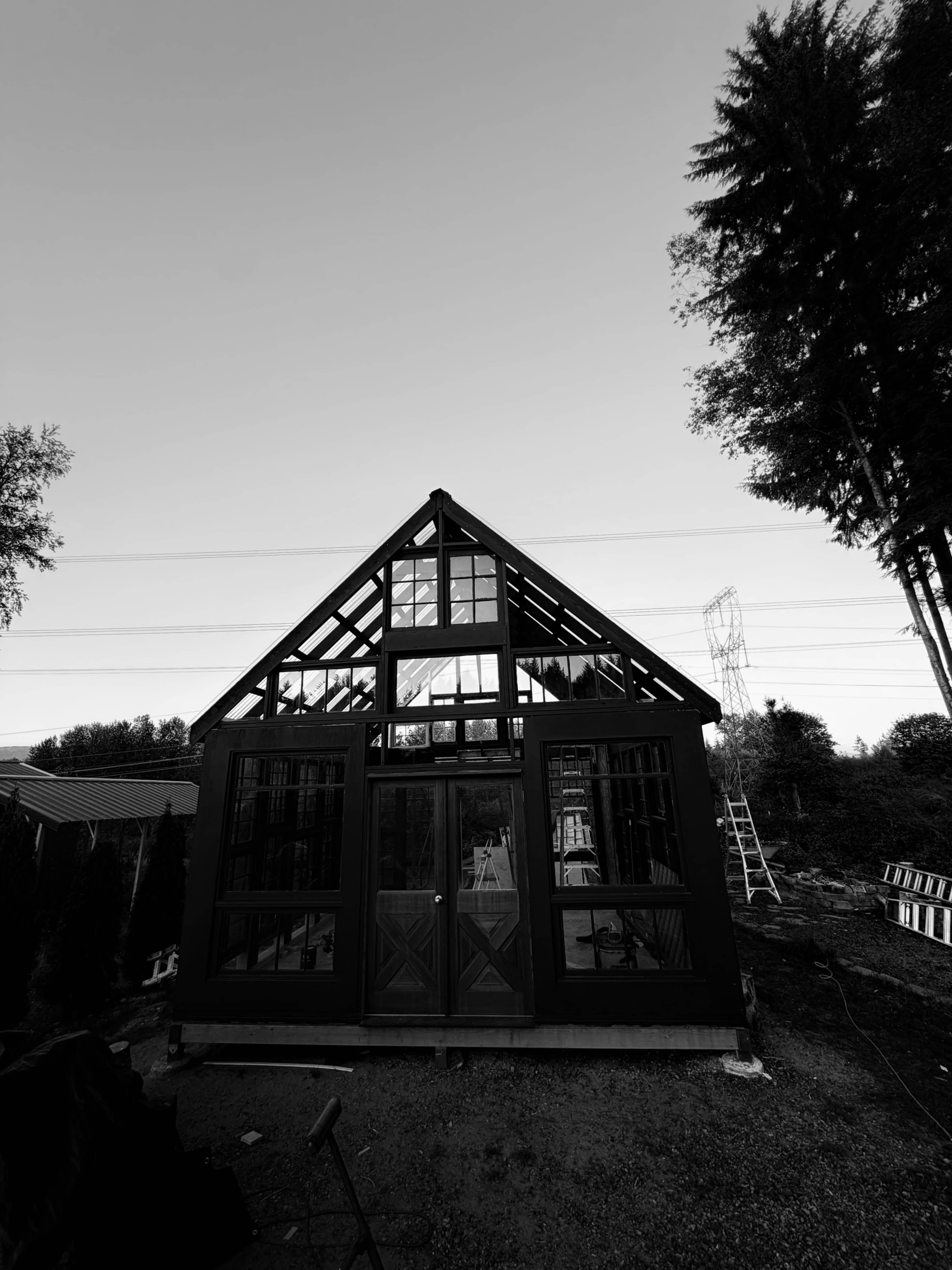 The image shows a partially constructed black and glass greenhouse with a triangular roof and large front windows, set against a twilight sky.