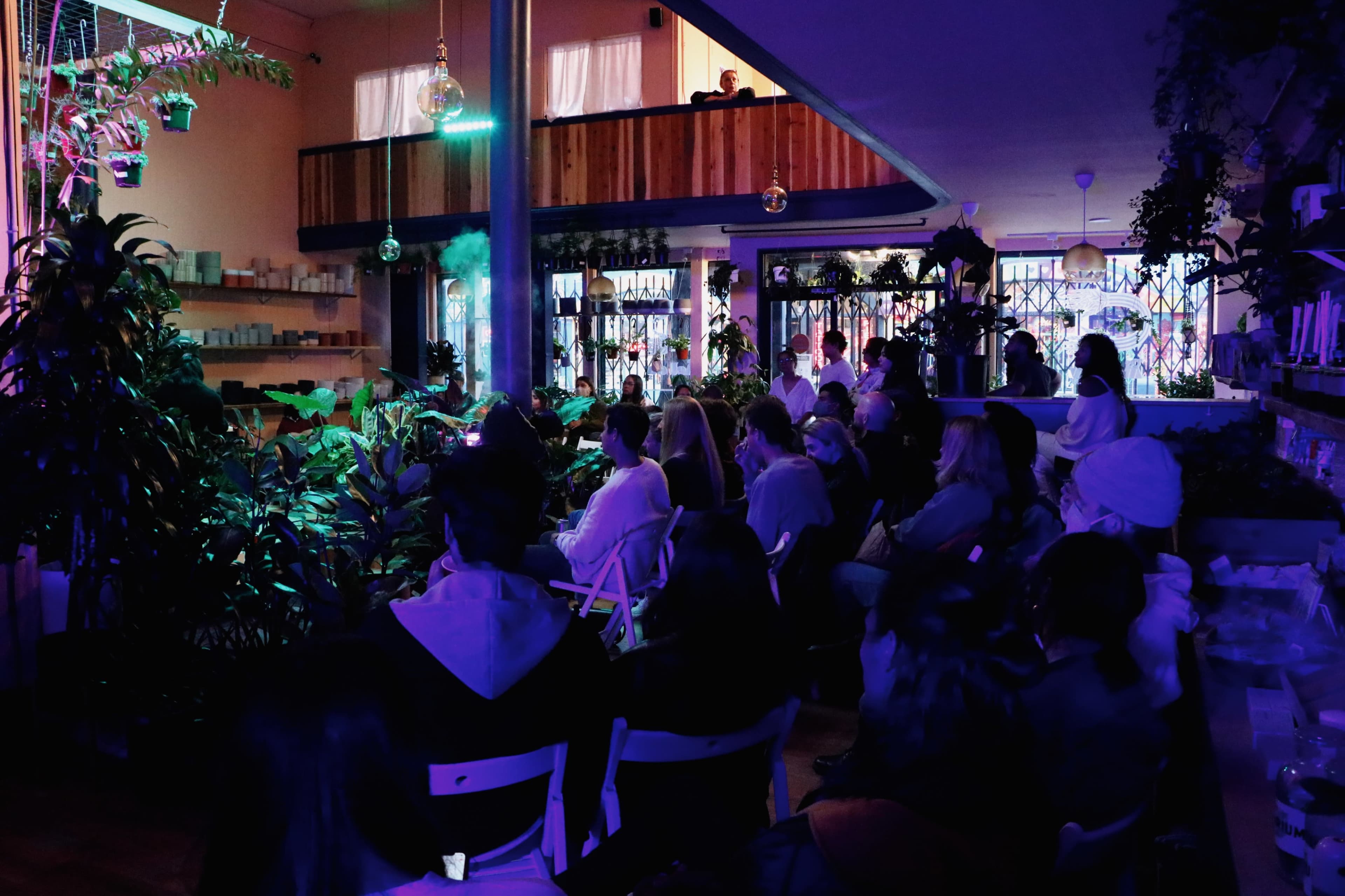 A group of people sits attentively in a dimly lit venue surrounded by plants, with colorful lights casting a glow on the scene.