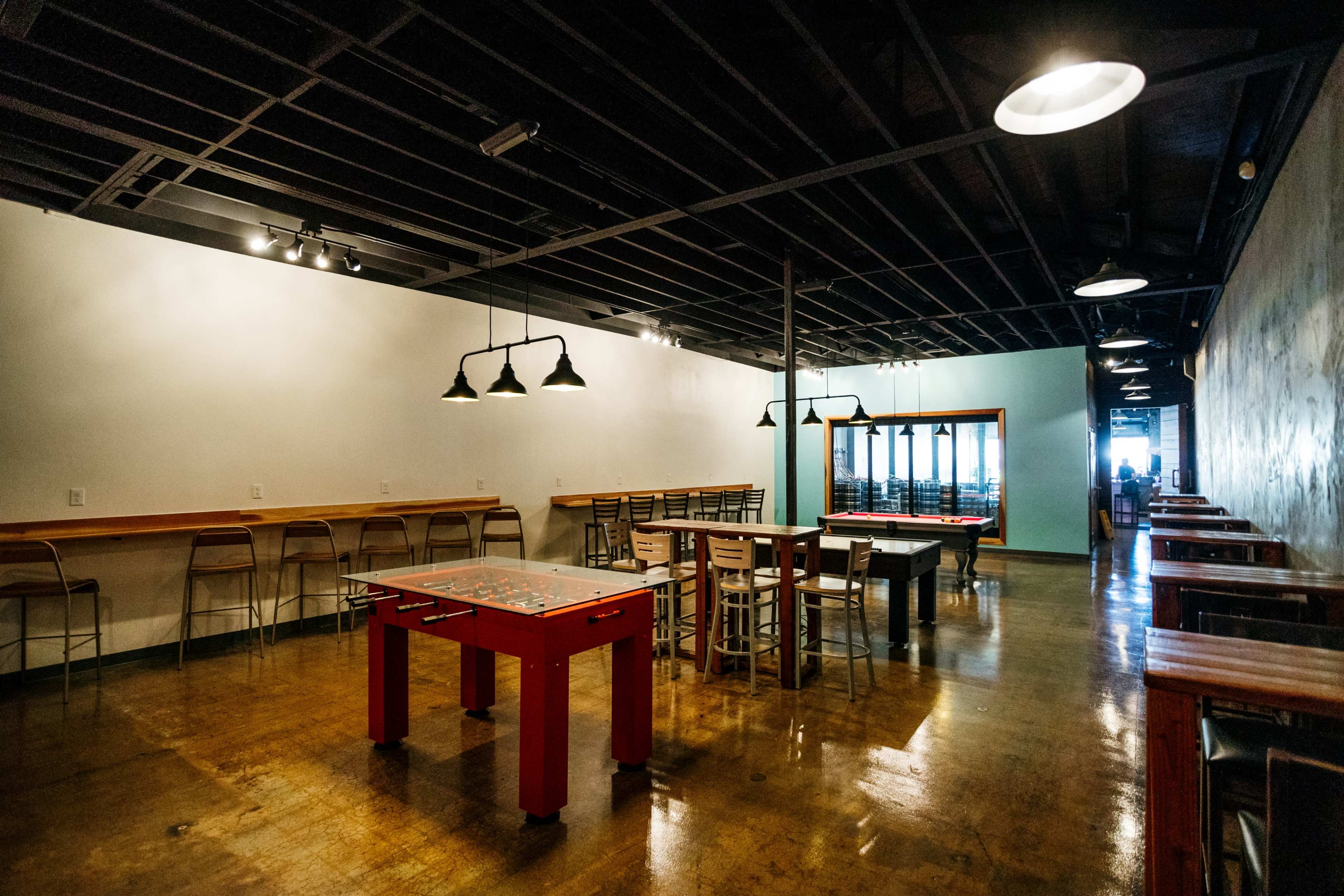 The image shows an empty game room featuring a foosball table and several tables and chairs along the walls.