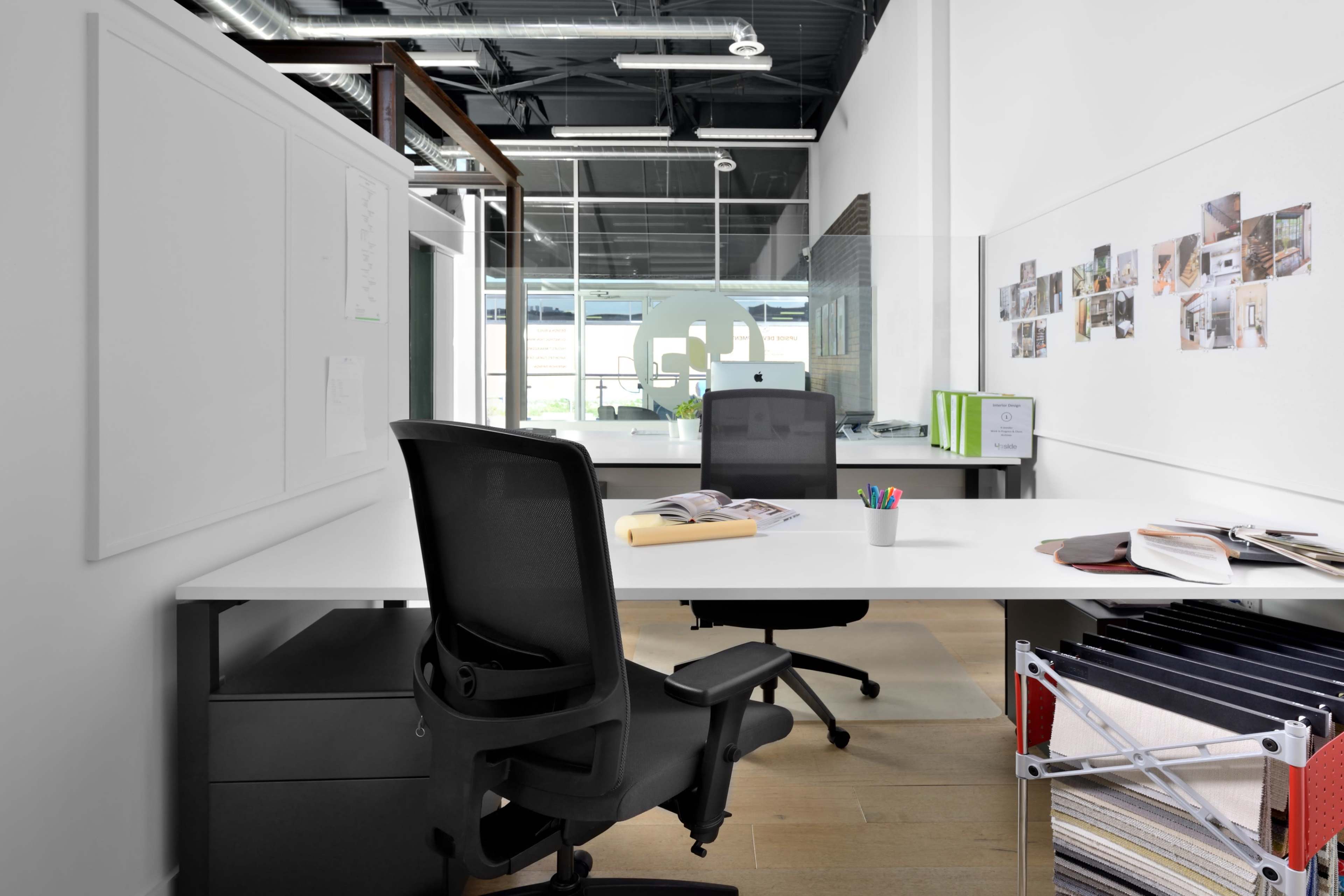 A modern office workspace featuring two desks, ergonomic chairs, and a wall with pinned photographs.
