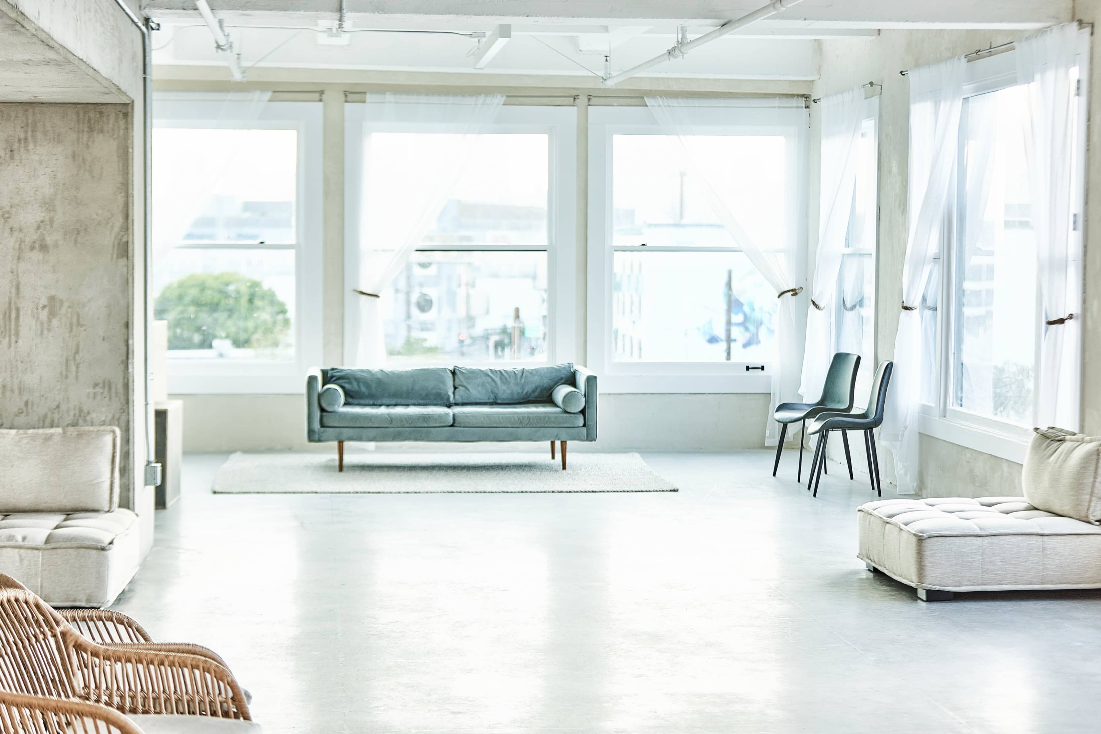 A spacious, well-lit room features a light blue sofa, a few chairs, and large windows with sheer curtains, set against a minimalist background.