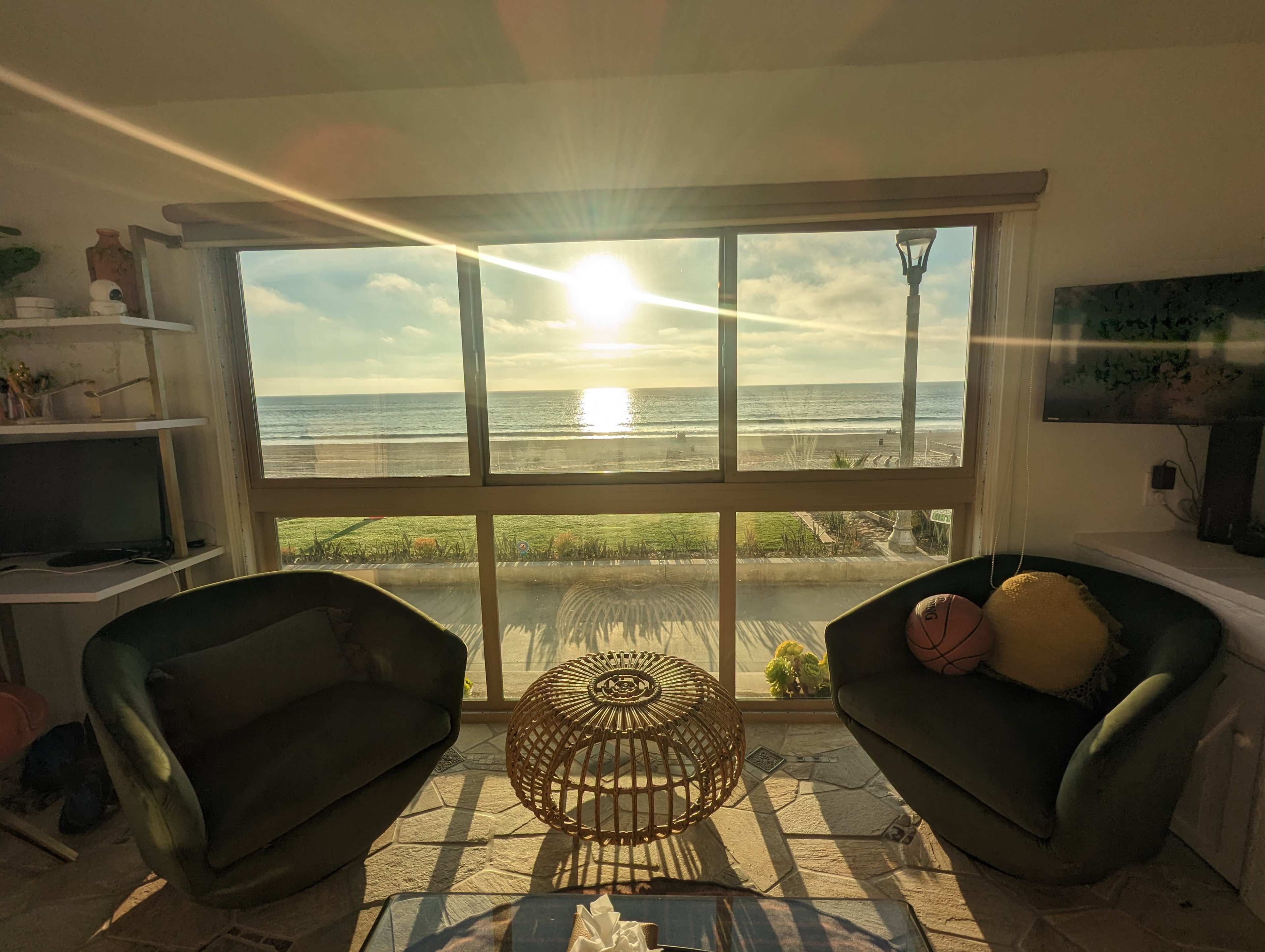 A cozy room with two green chairs and a view of the ocean under a setting sun, visible through a large window.