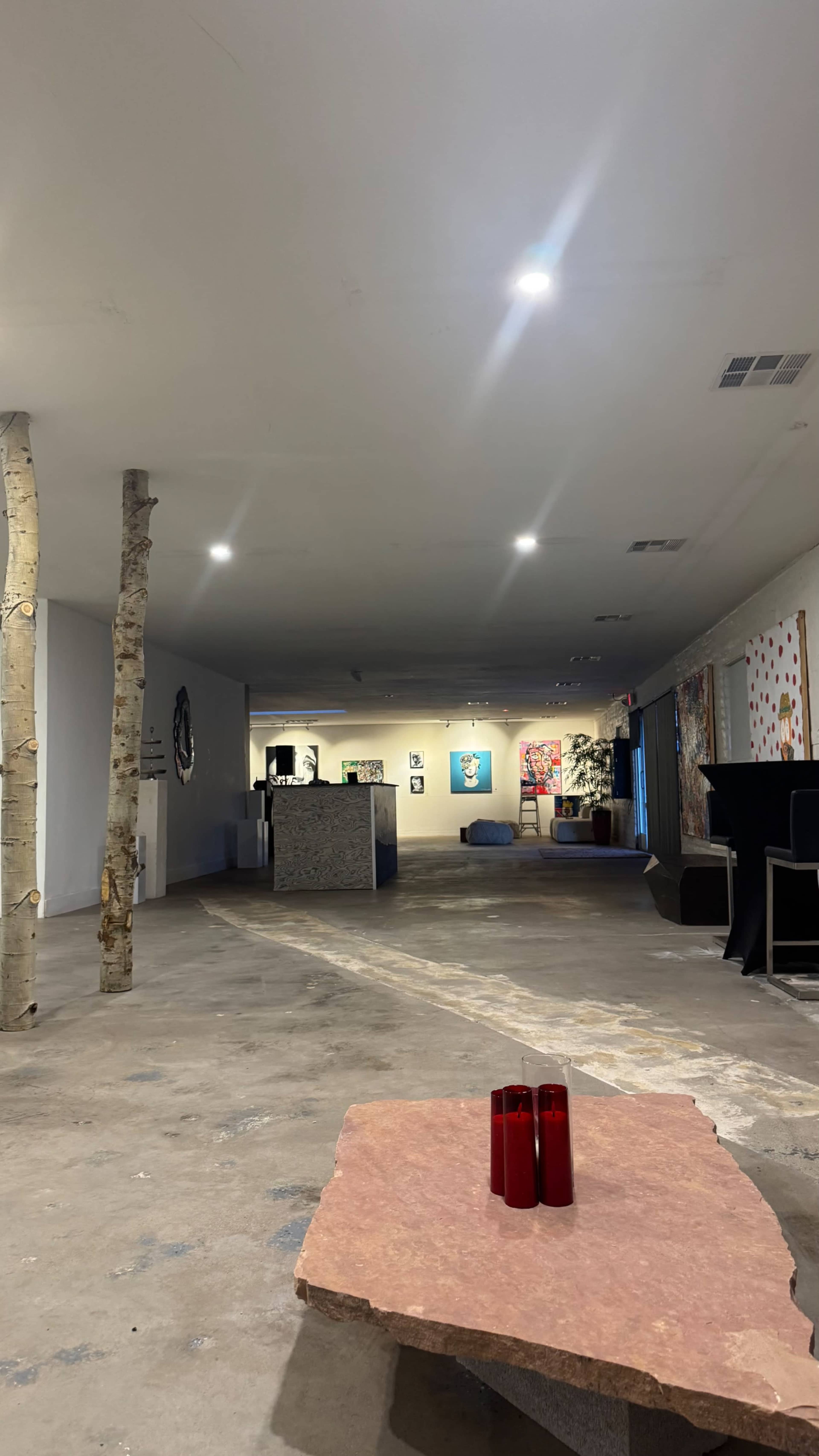 An expansive, contemporary art gallery features concrete flooring, sparse furniture, and various artworks displayed on the walls, with two columns of bare trees standing prominently in the center.