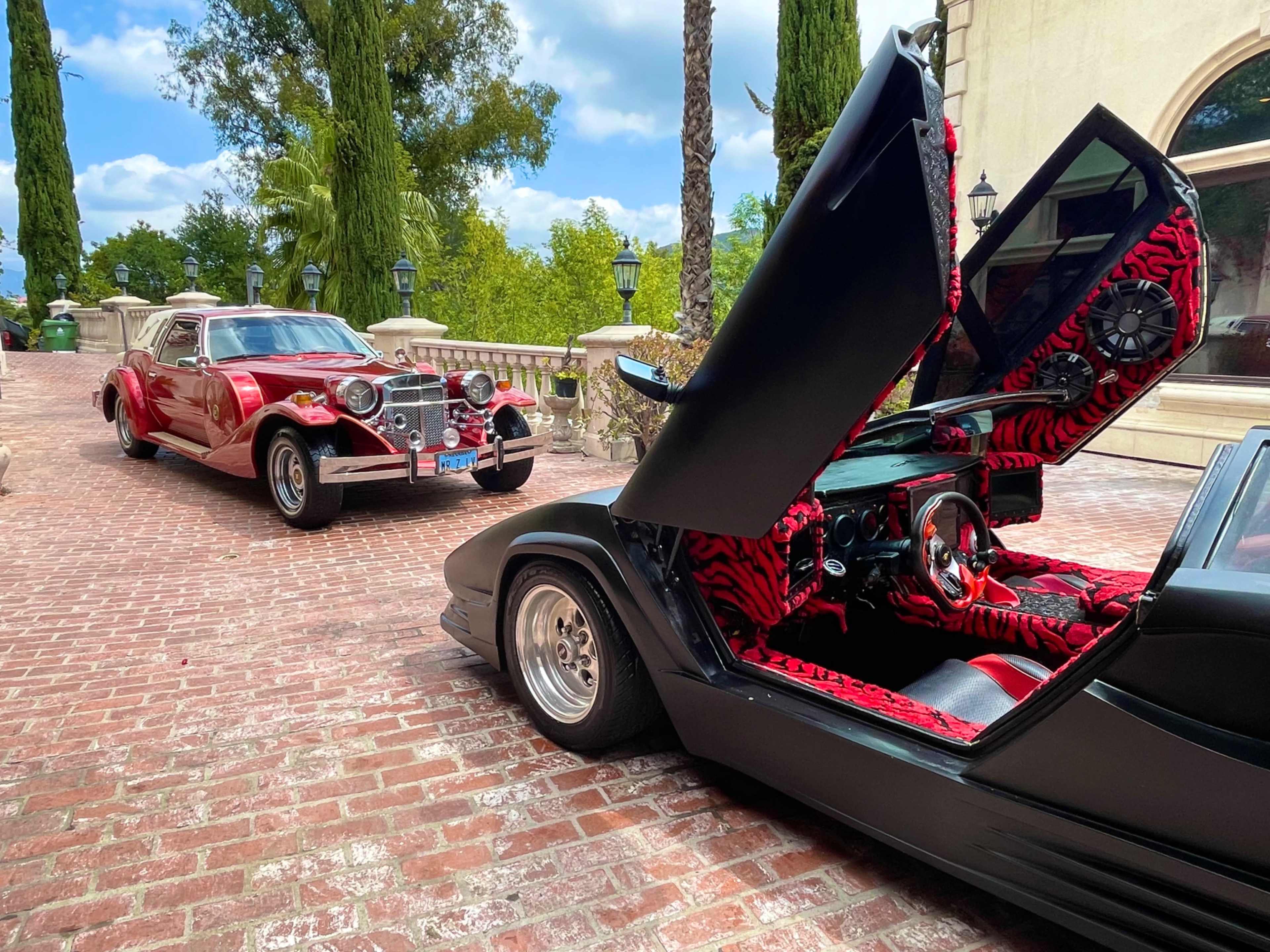 Two classic cars, one with its doors open and an interior featuring red and black stripes, are parked on a brick driveway in a landscaped area.
