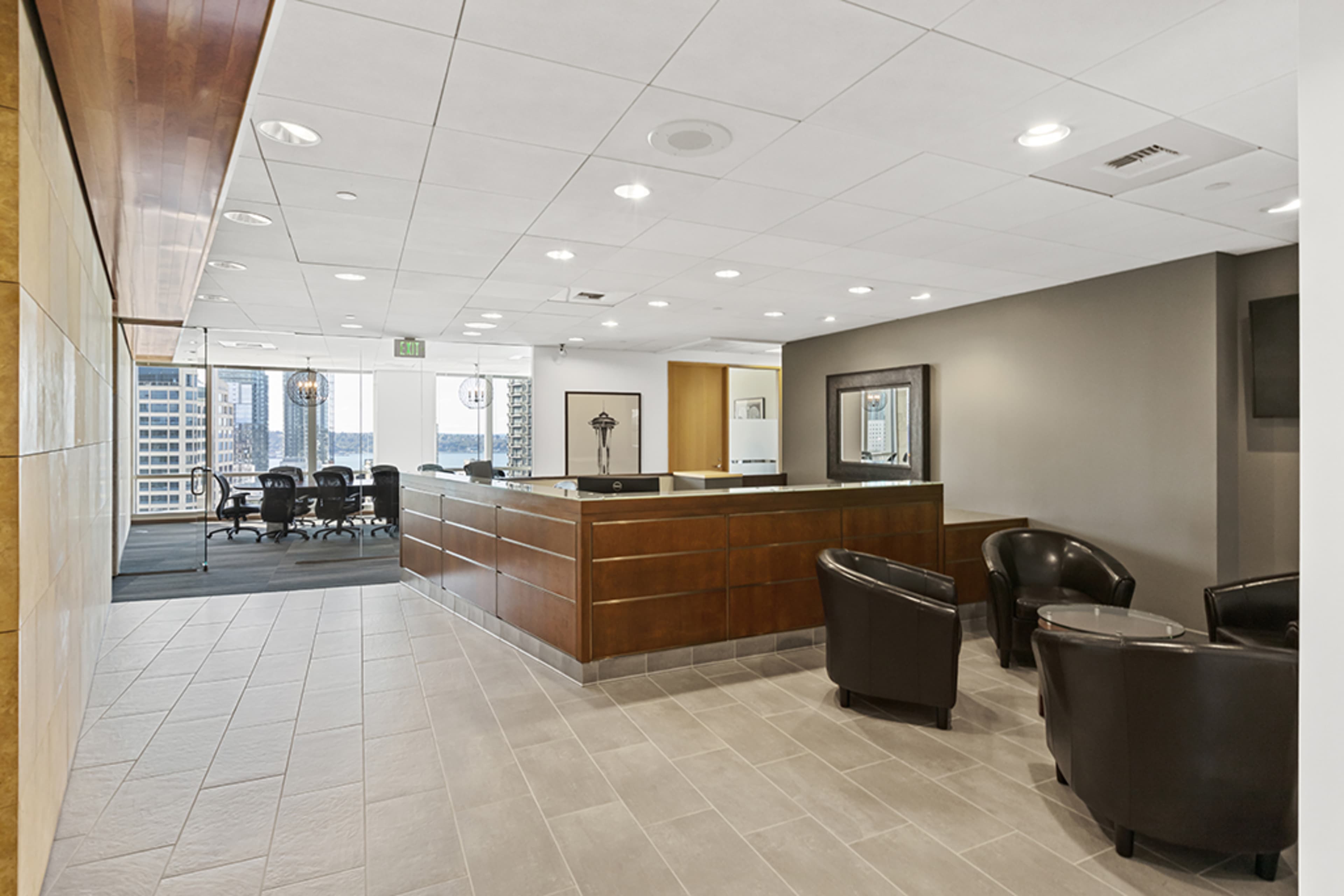 A modern office reception area with a wooden desk, seating for visitors, and a meeting room visible in the background.