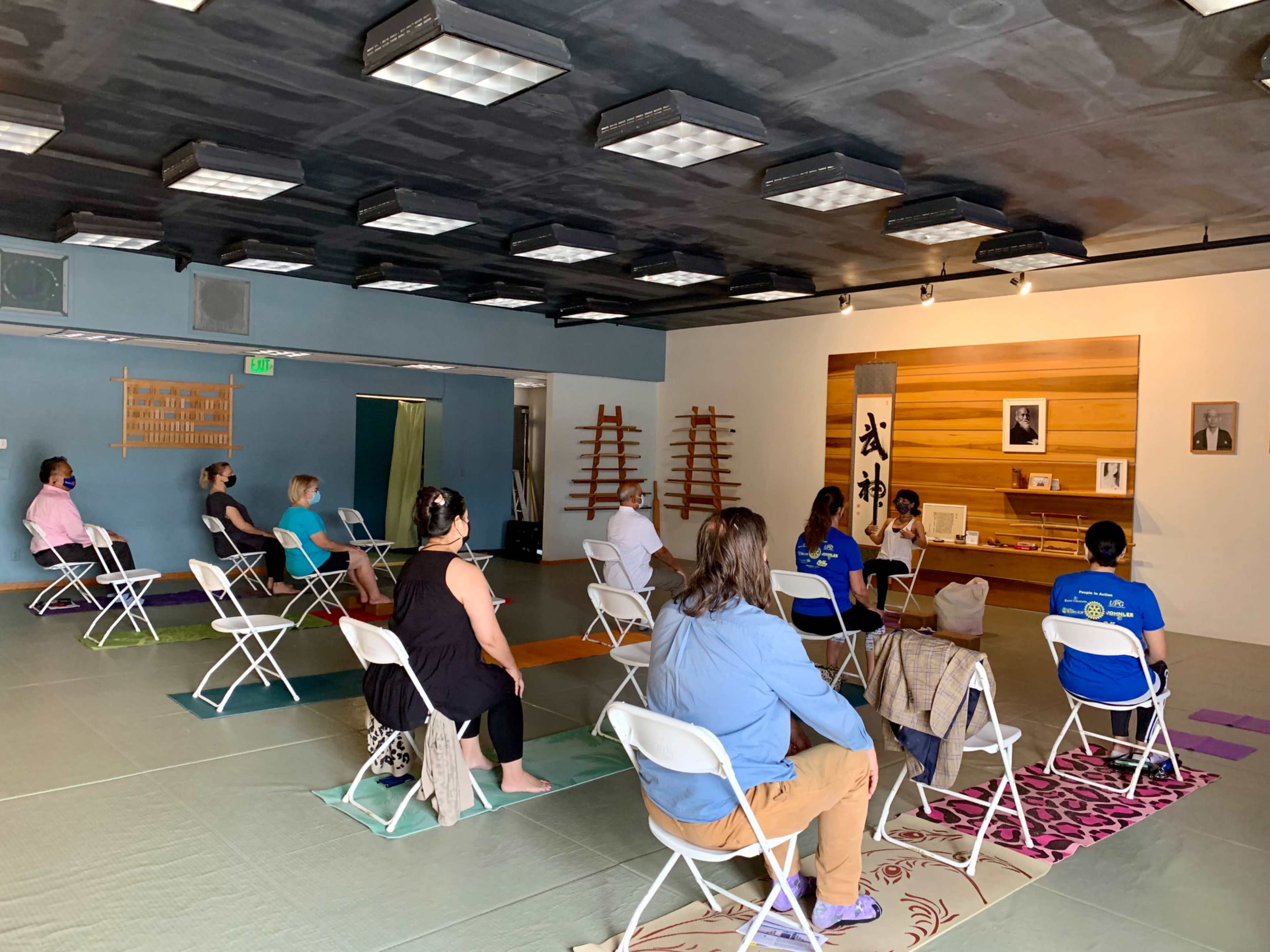 A group of people sits on folding chairs in a spacious room with tatami mats, facing a small altar and a wall displaying an image and text.