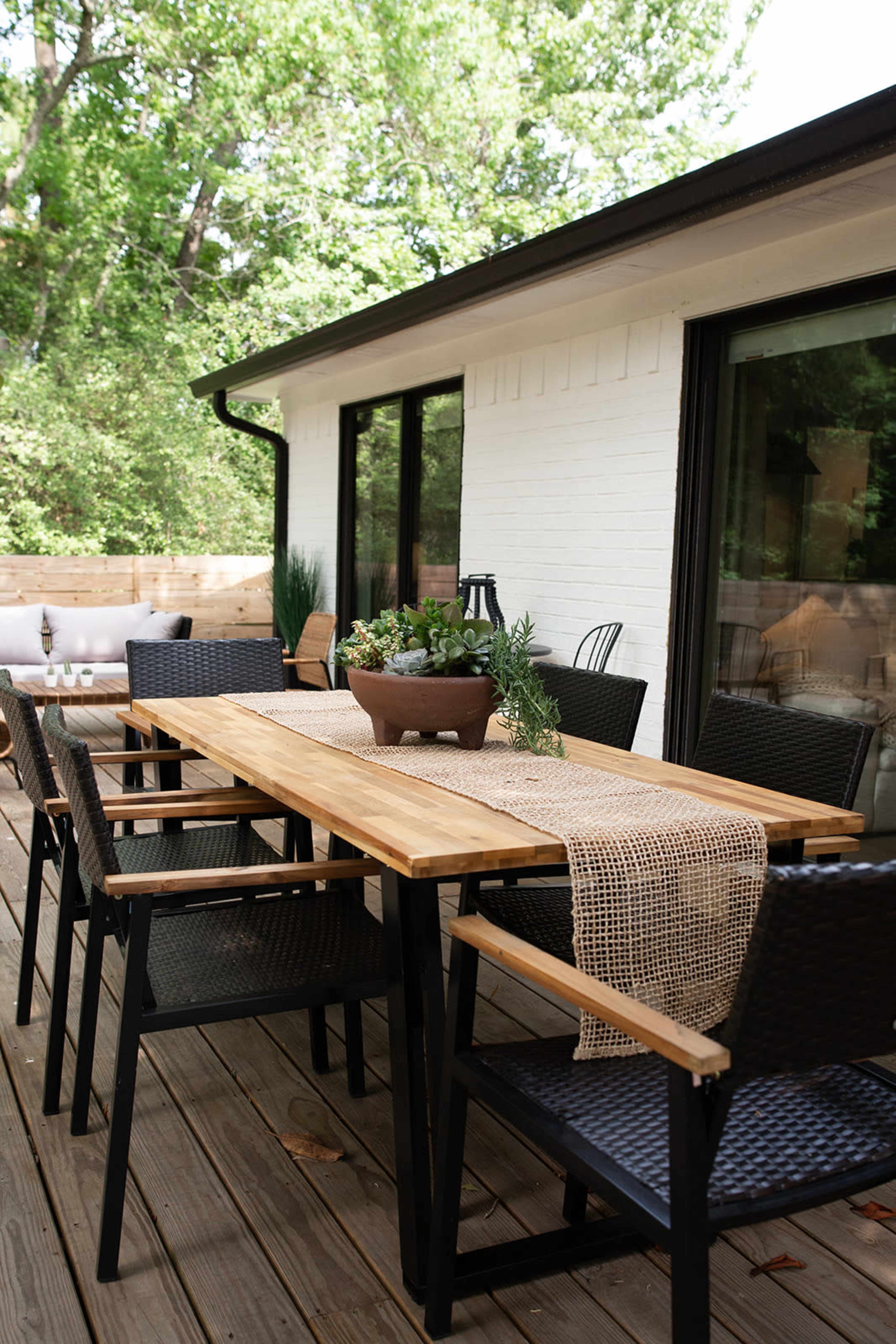 A wooden dining table with black chairs is set on a wooden deck, surrounded by greenery and adjacent to a modern home.