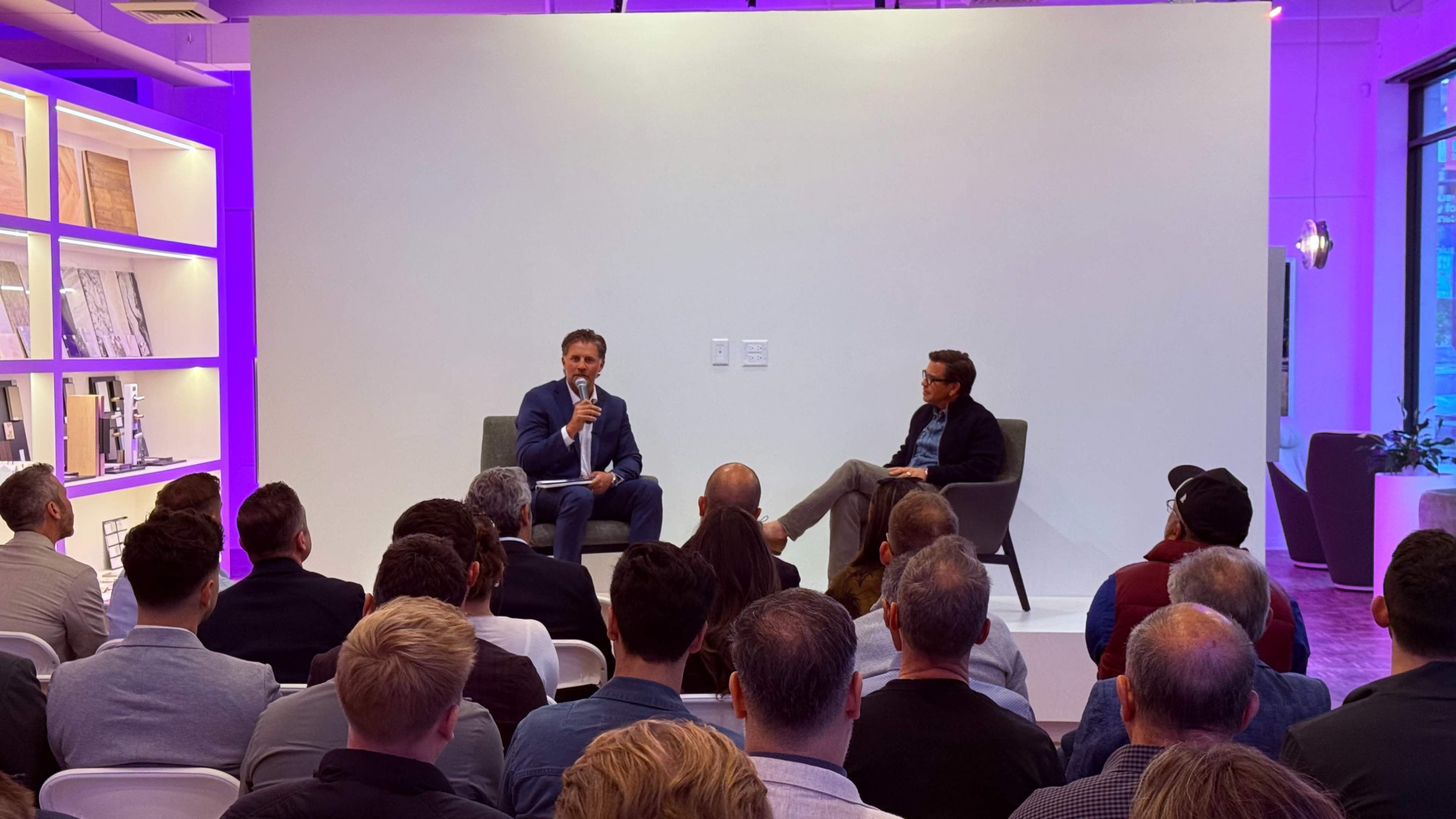 A speaker addresses an audience during a discussion event while seated on a stage with another participant.