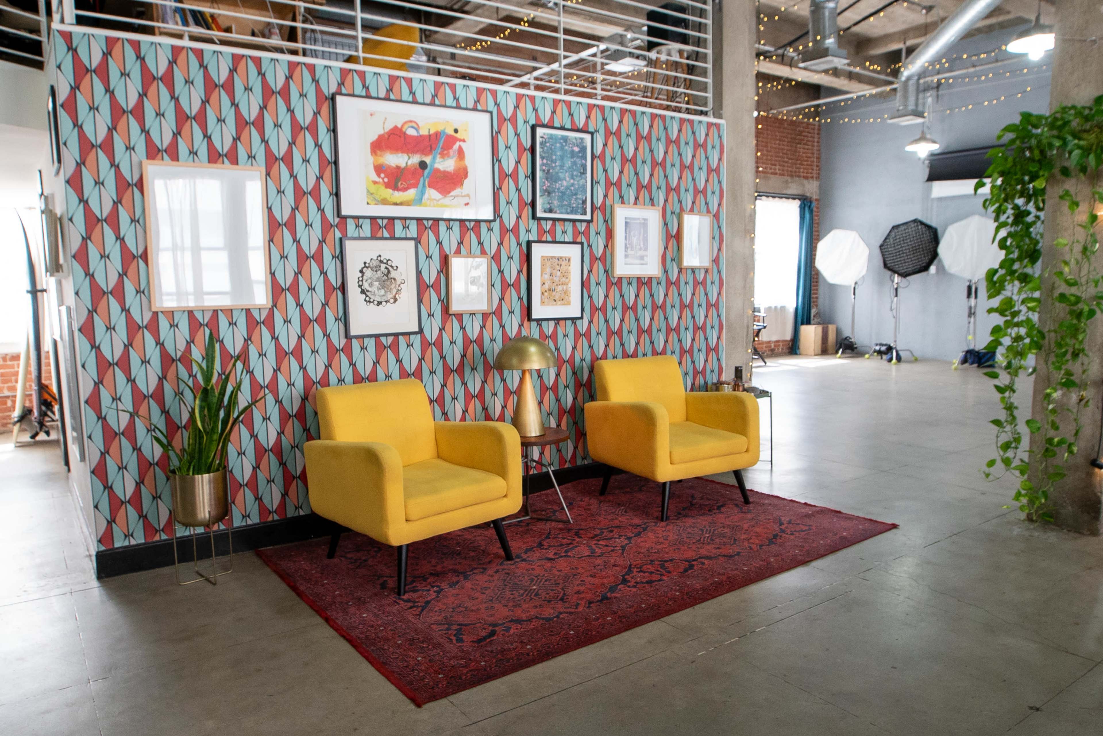 A stylish interior with two yellow chairs positioned in front of a patterned wall, decorated with framed artwork and a potted plant, on a red area rug.