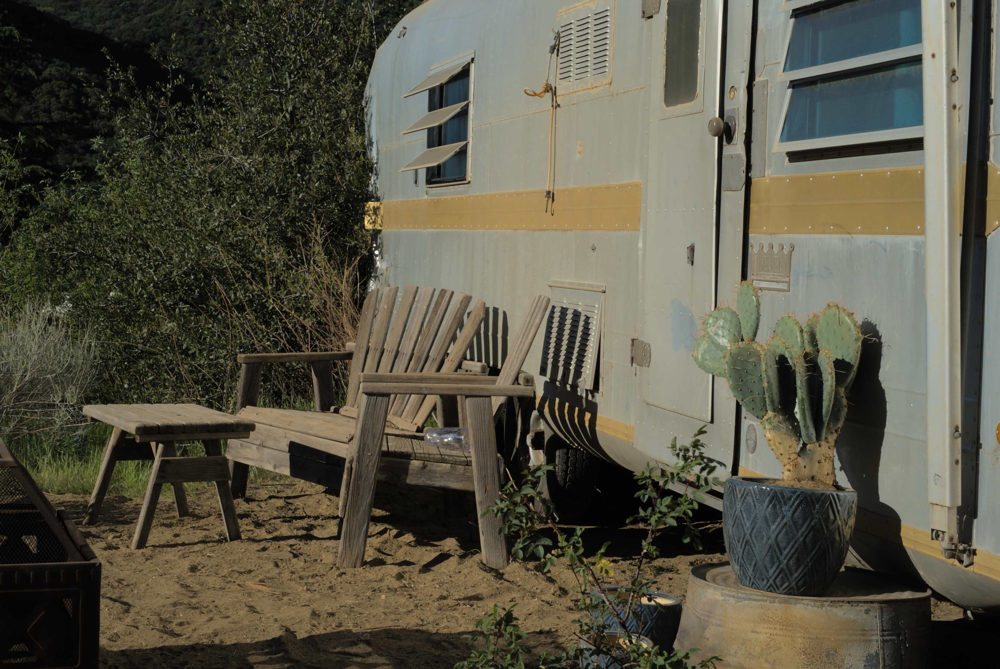 A vintage trailer is parked in a sandy area, accompanied by wooden seating and a potted cactus.