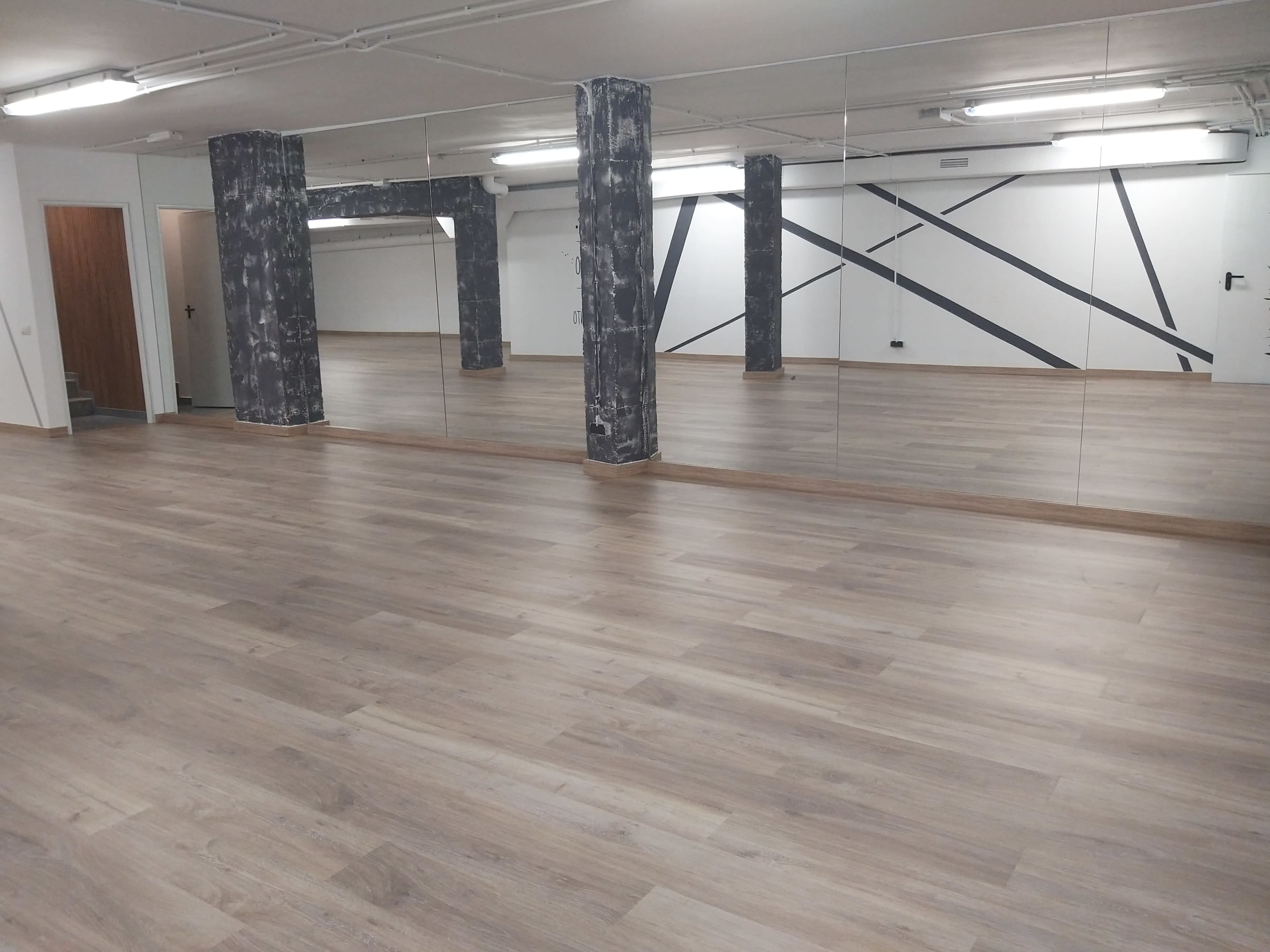 A spacious room with wooden flooring, mirrored walls, and exposed structural columns featuring a textured design.