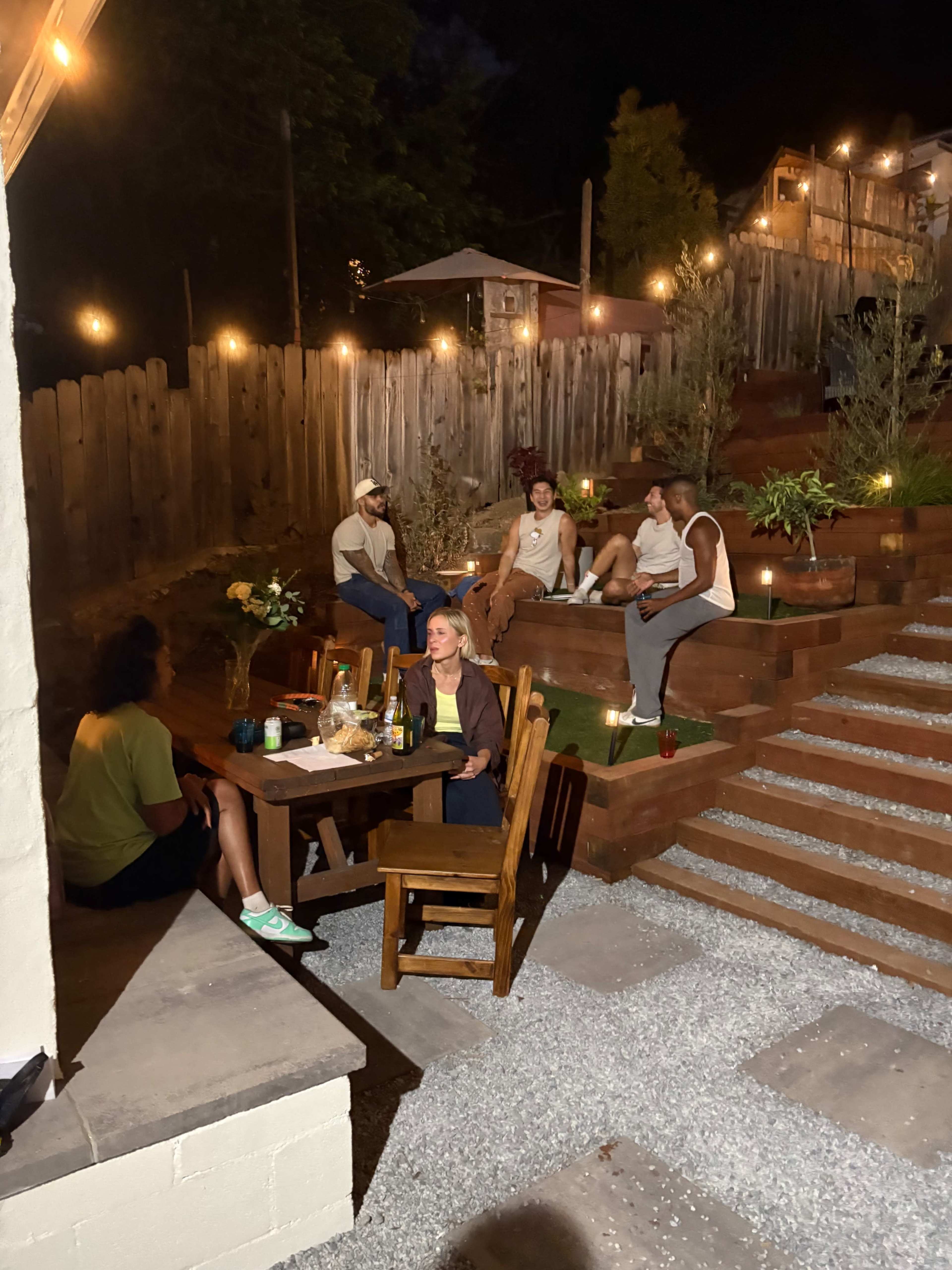 A group of five people is seated around tables in a backyard, illuminated by string lights, enjoying drinks and conversation.