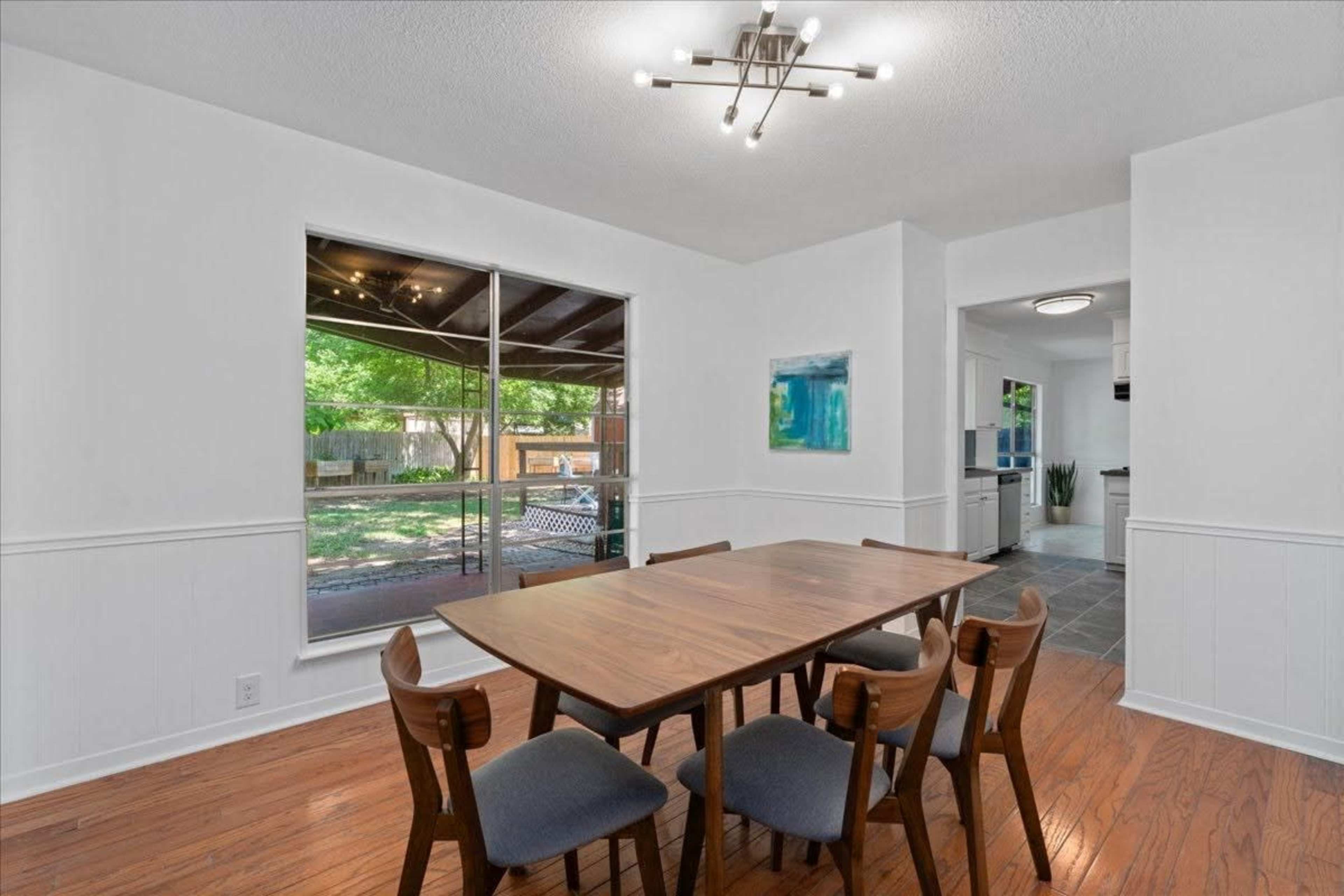 A modern dining area features a wooden table surrounded by chairs, with large windows offering a view of a backyard.