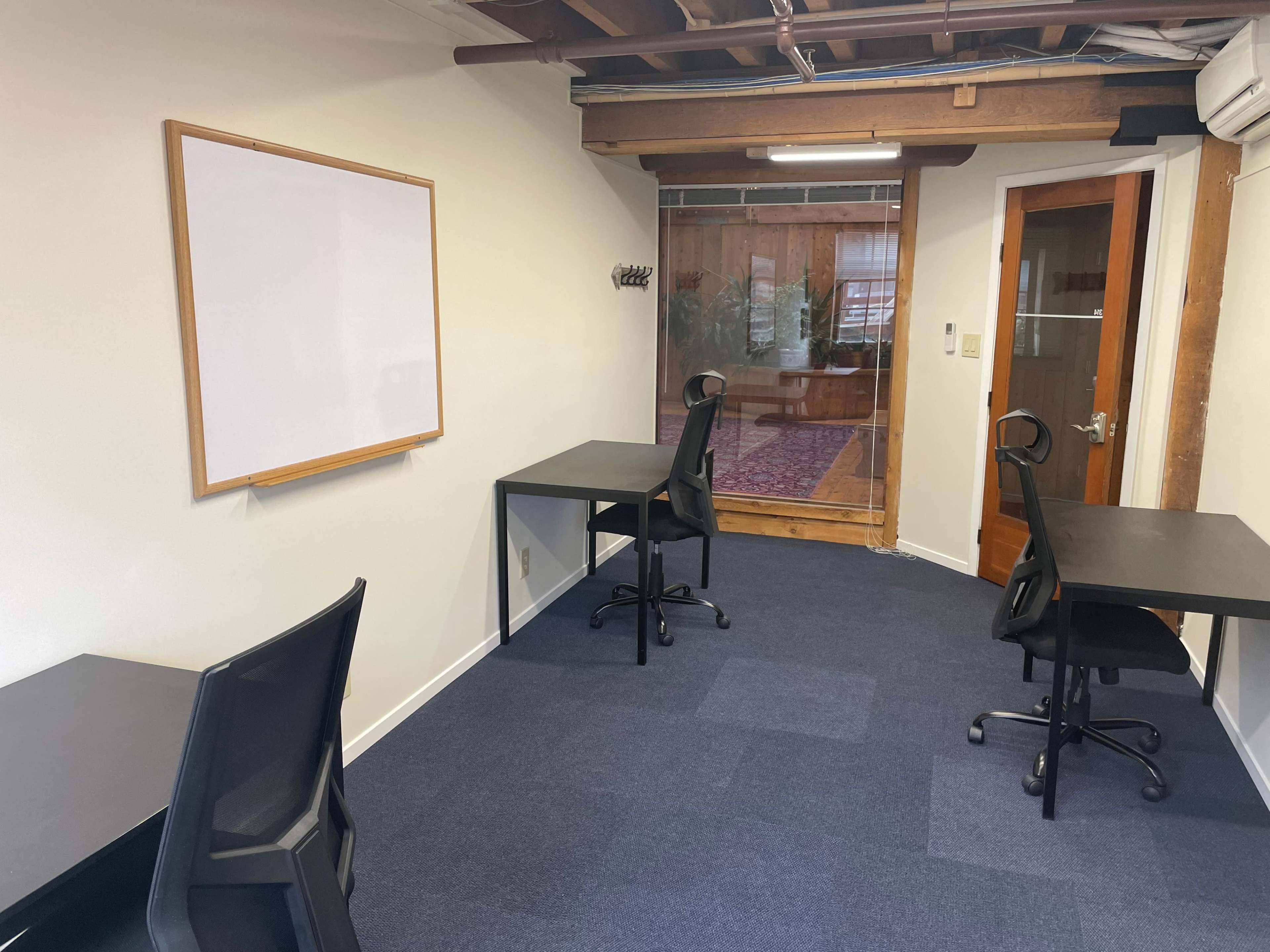 The image shows a small office space with two black desks and chairs, a whiteboard on the wall, and a door leading to another room.