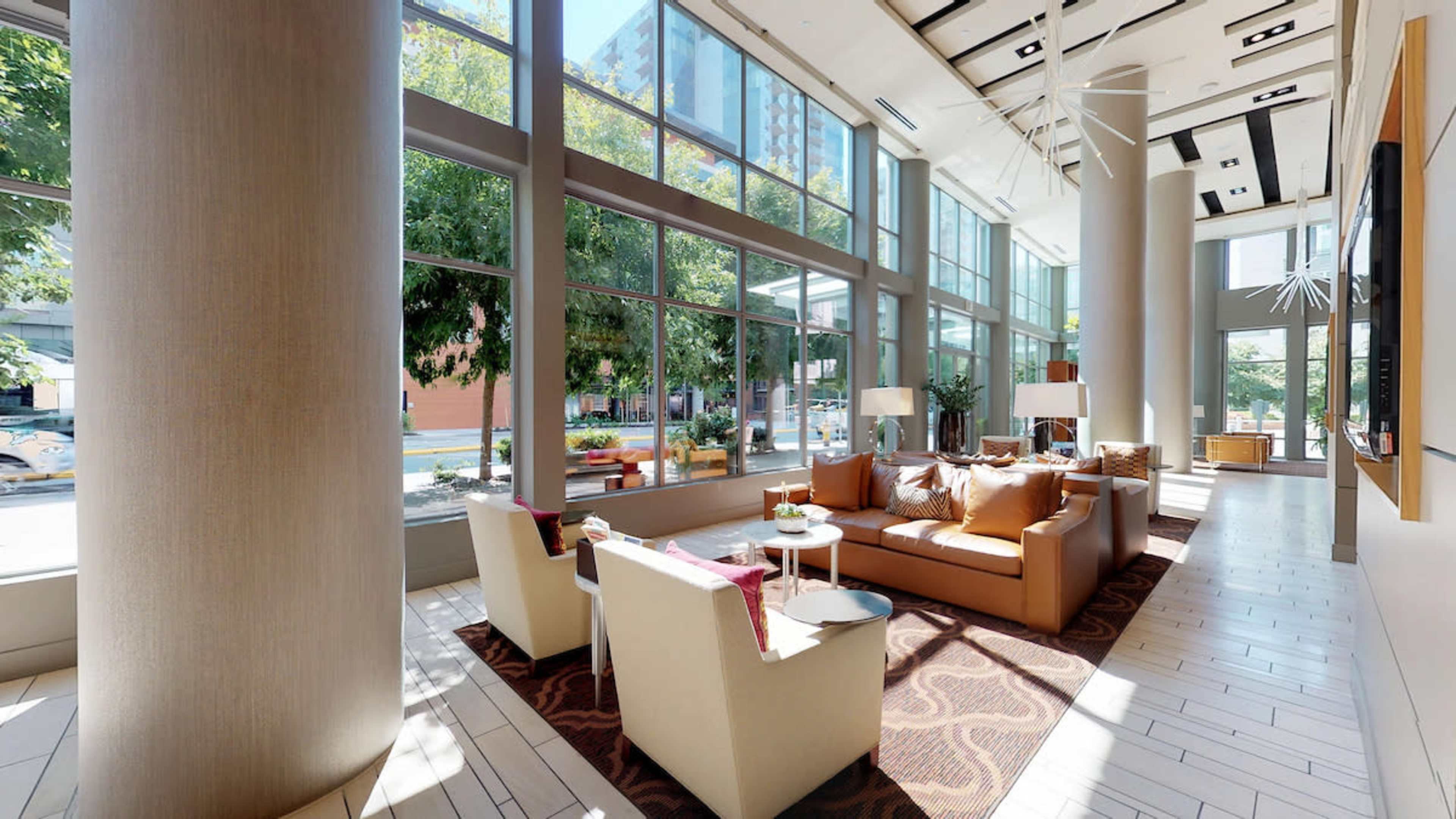 The image shows a modern lobby area with large windows, comfortable seating, and decorative plants.