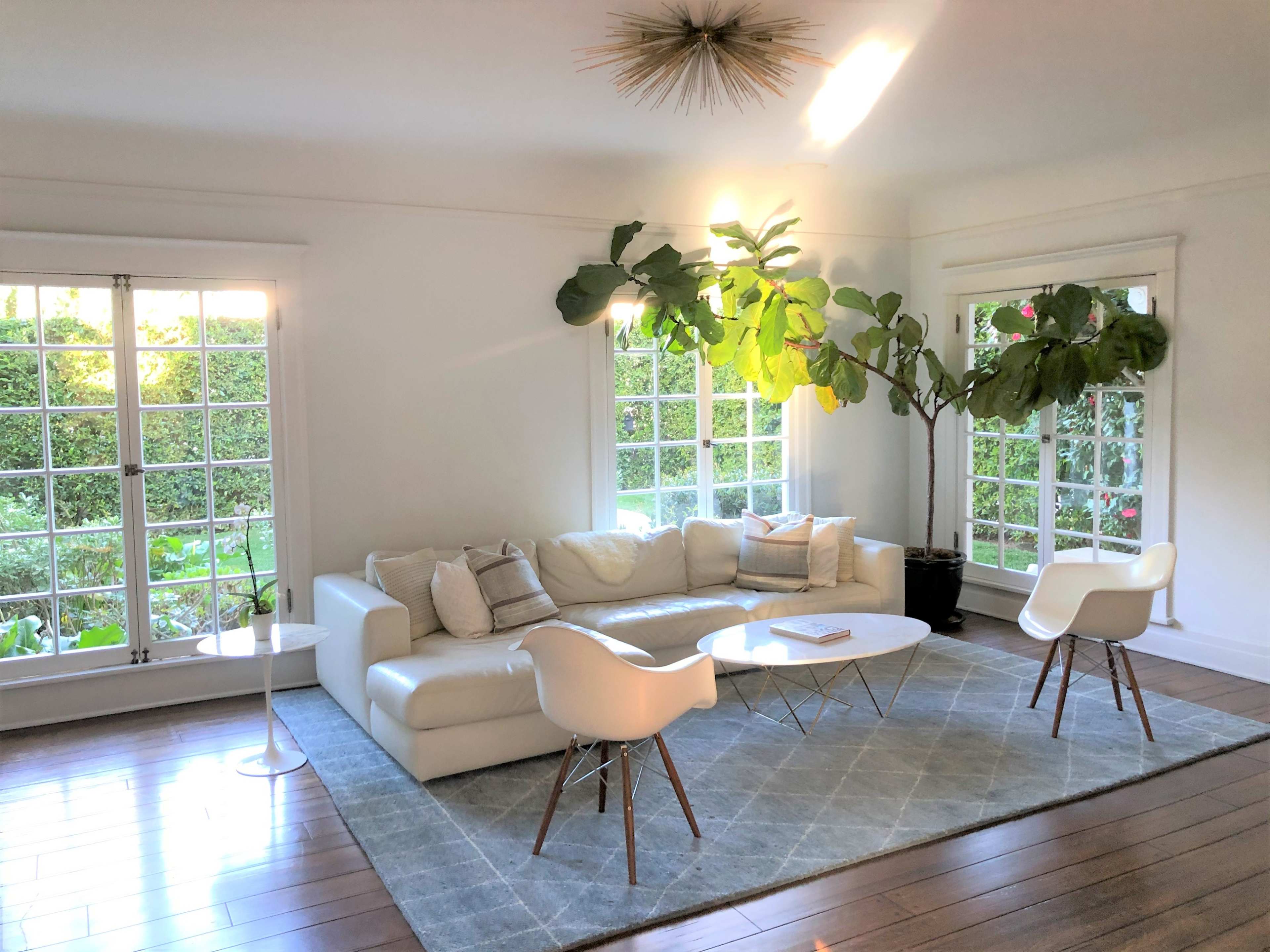 A modern living room featuring a white sectional sofa, two white chairs, a round coffee table, and large windows with green foliage outside.