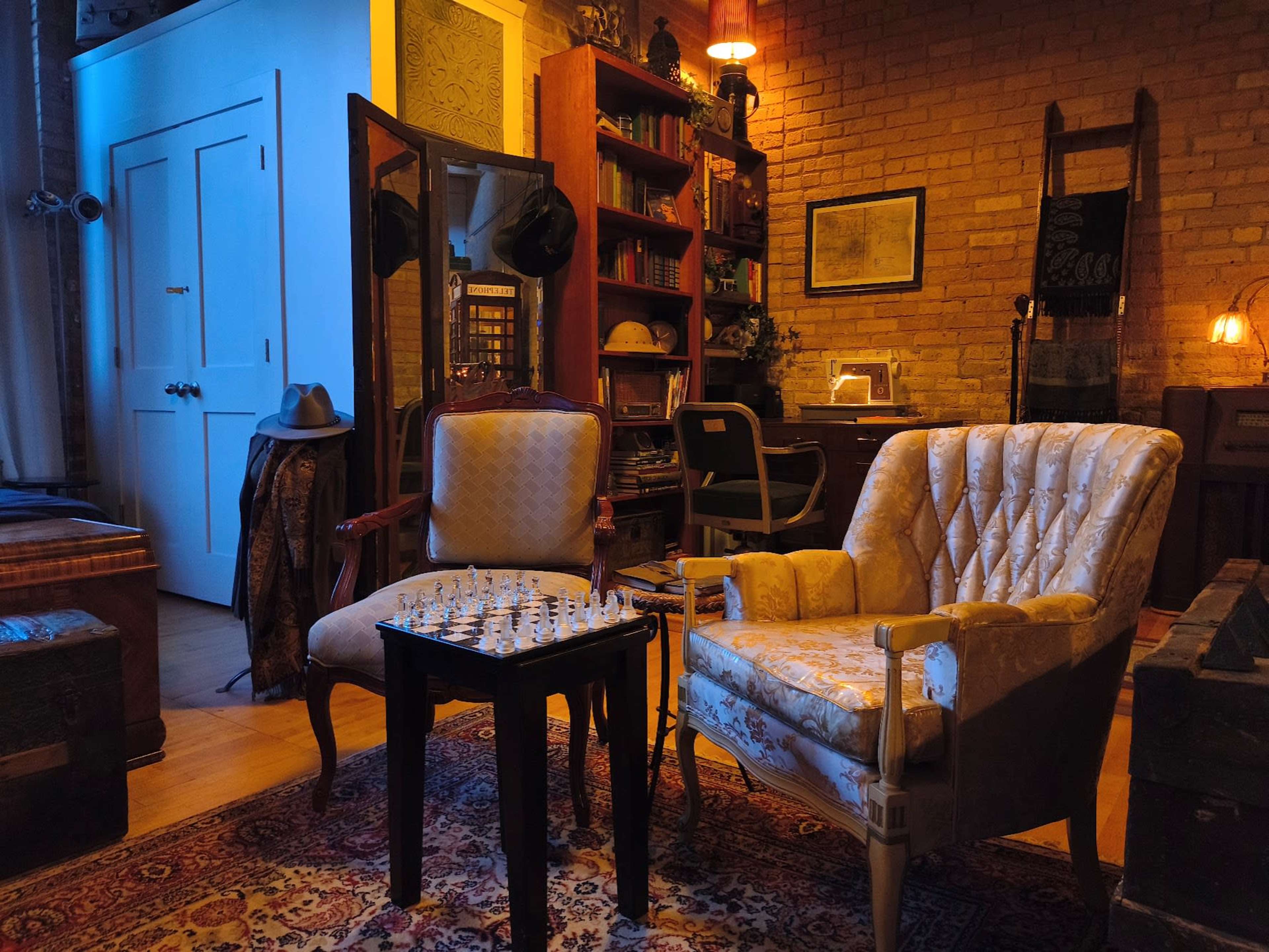 The image features a cozy room with two vintage armchairs facing a small chess table, surrounded by bookshelves and warm lighting.