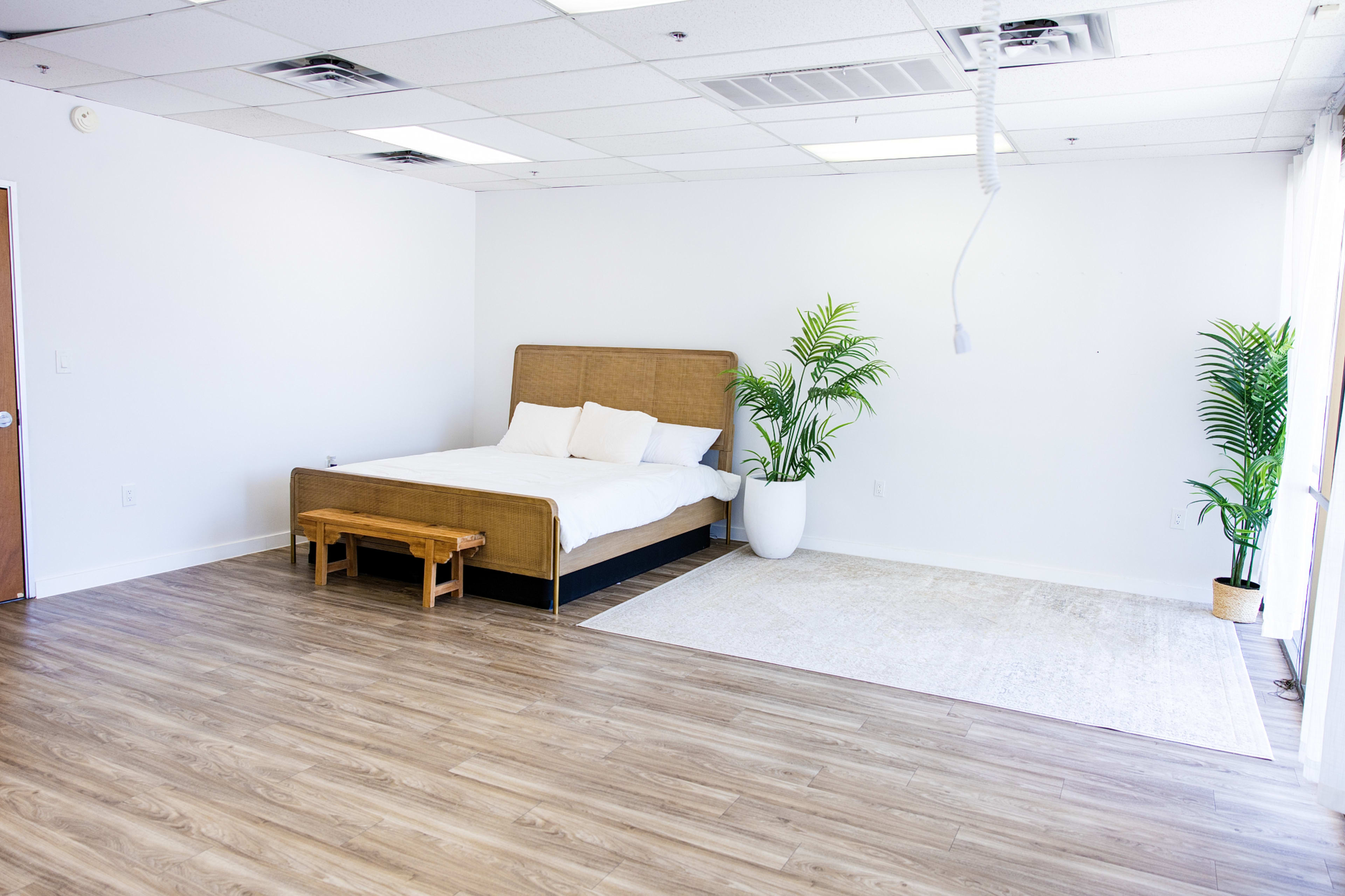 The image depicts a spacious, minimalist bedroom featuring a large bed with white linens, a wooden bench, and potted plants against a light-colored wall and floor.