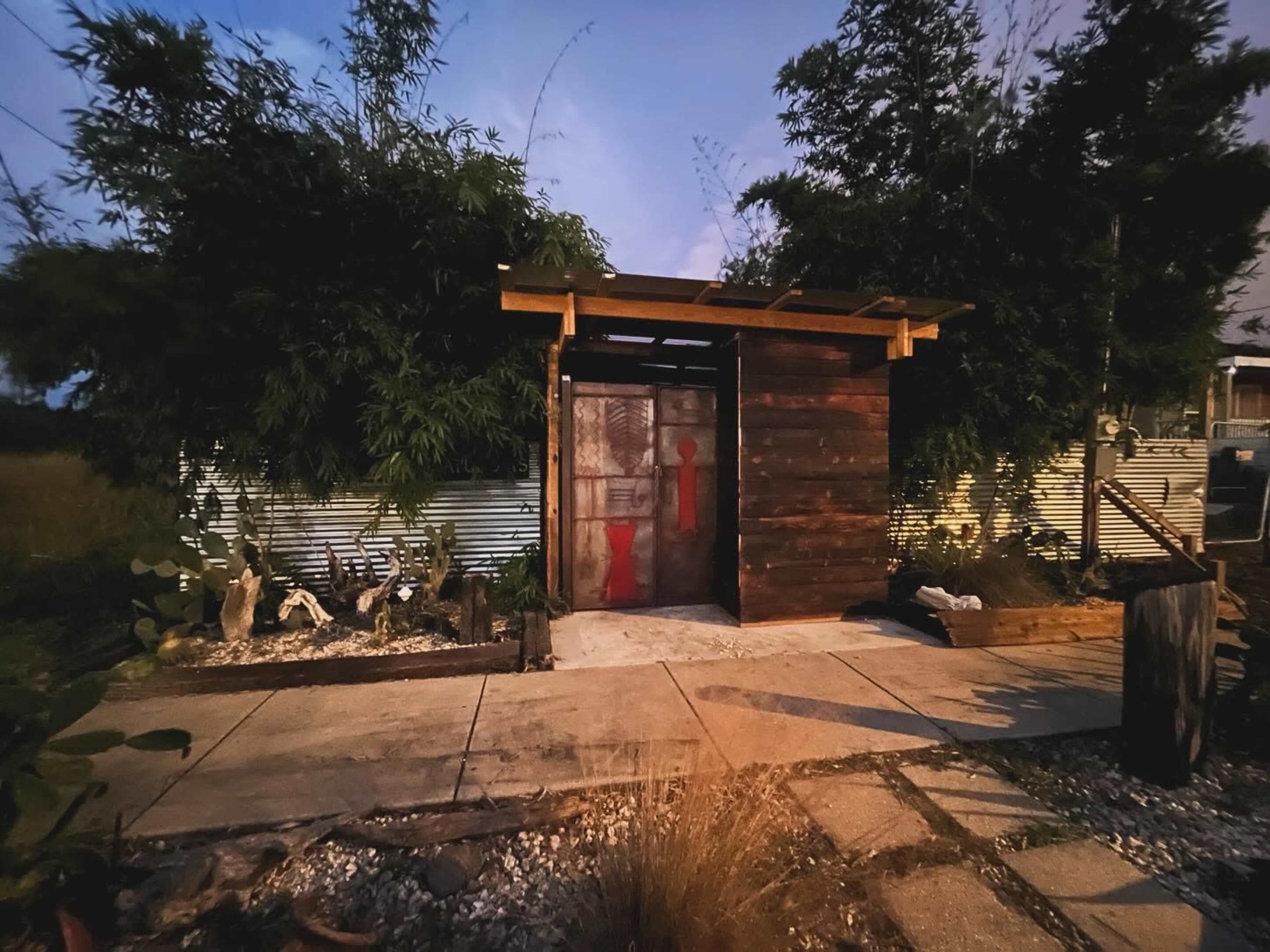 A small wooden structure with a rustic door is set against a backdrop of greenery and gravel pathways in low evening light.