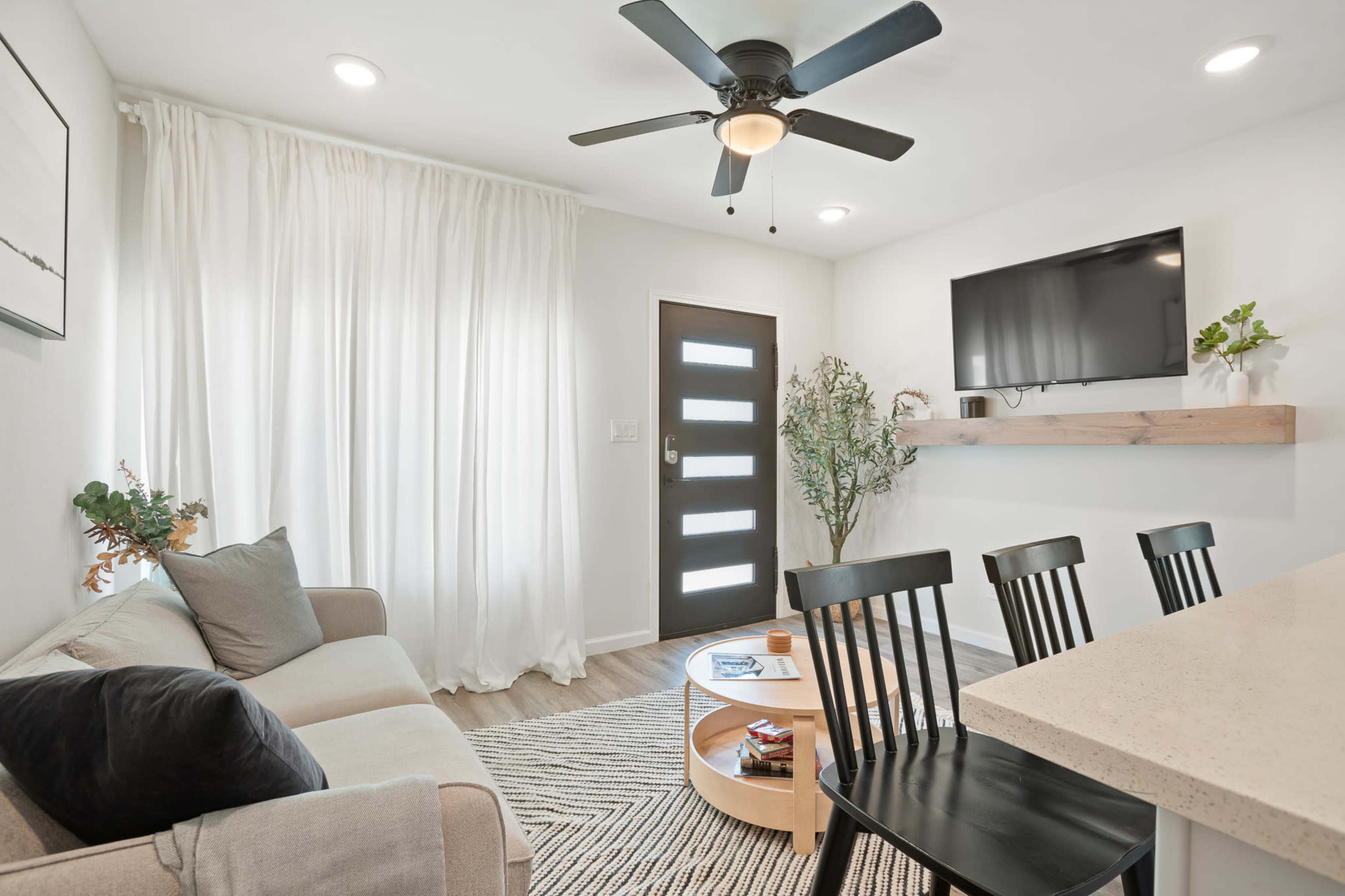 The image shows a modern living space featuring a light-colored sofa, a circular coffee table, a flat-screen TV mounted on the wall, and a door with vertical windows beside it.