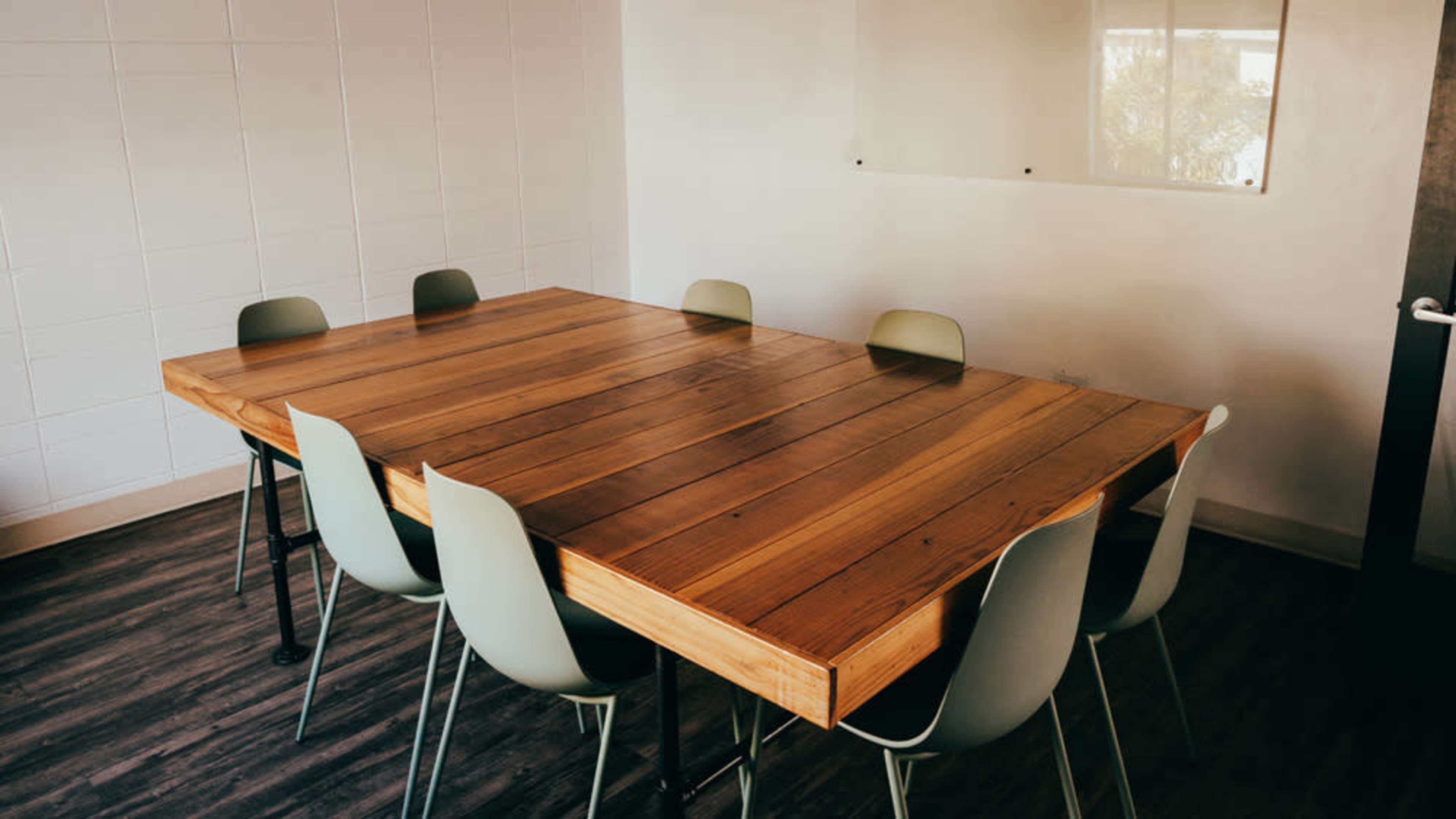 A large wooden table with eight chairs is placed in a minimalistic meeting room with white walls and a glass board.