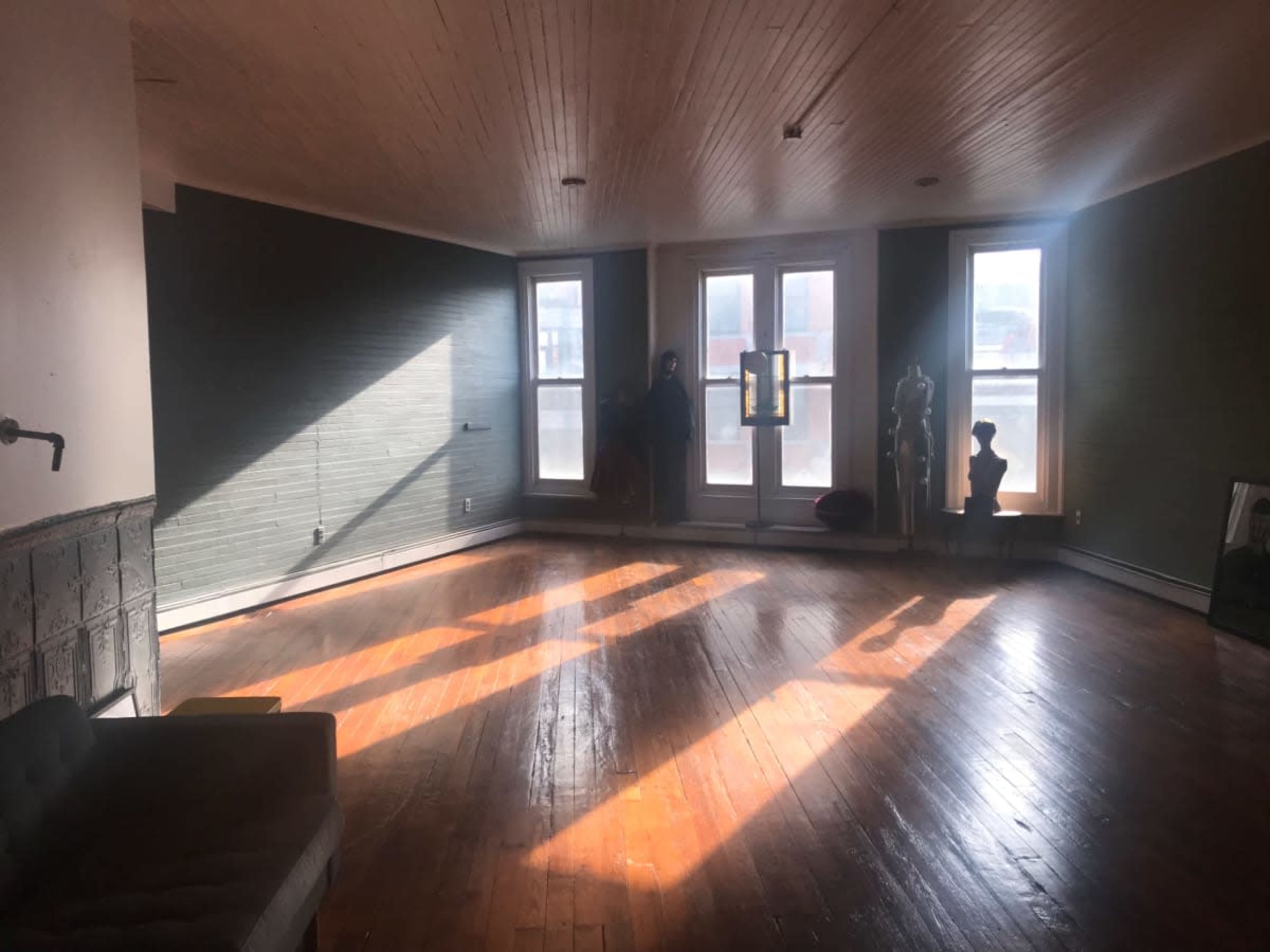 A spacious, sunlit room features large windows casting shadows on the wooden floor and a few scattered furniture pieces.