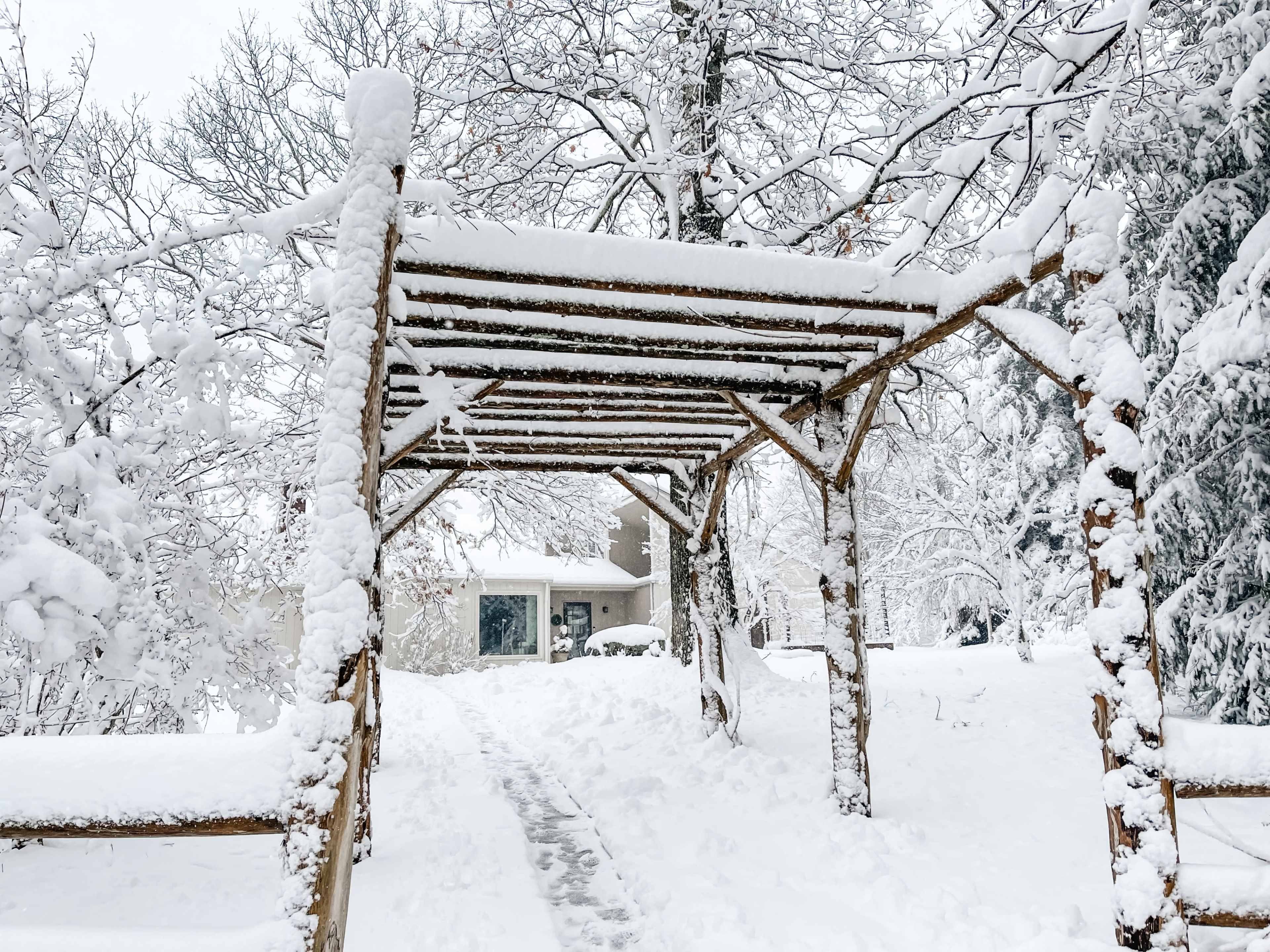 A wooden pergola covered in snow stands in a snowy landscape leading to a house in the background.