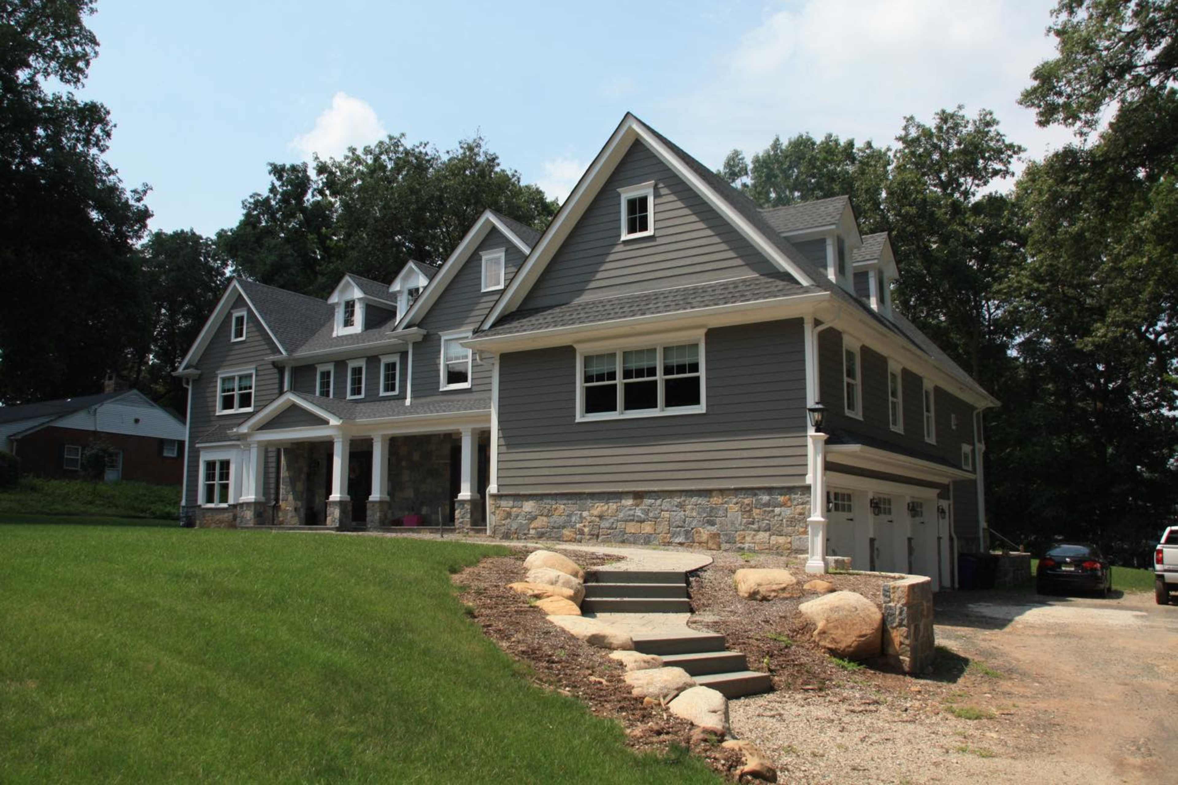 The image shows a large, multi-story gray house with stone accents and a front lawn, situated on a grassy lot.