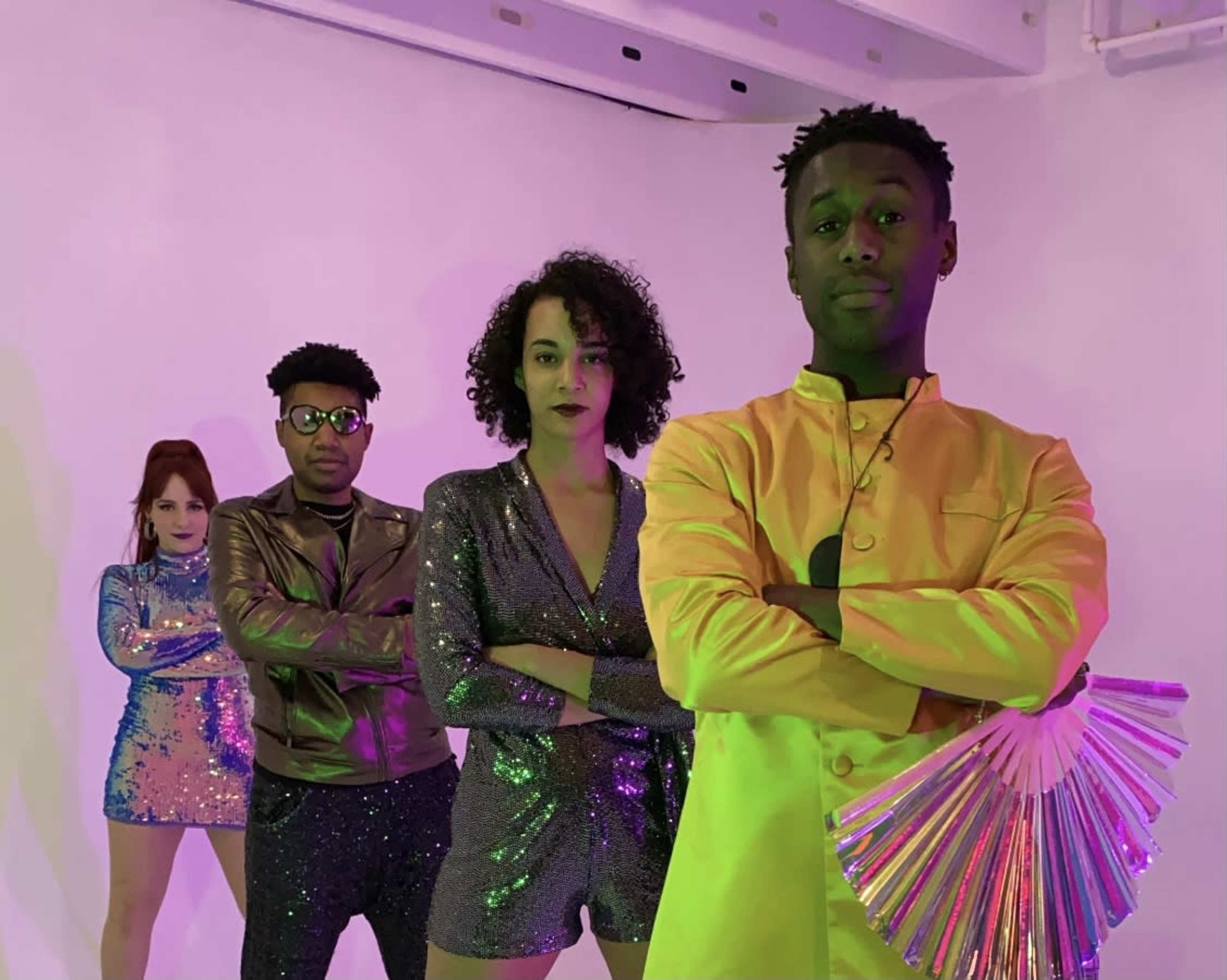 Four people pose in a brightly lit studio wearing colorful, sequined outfits, with one holding a fan.