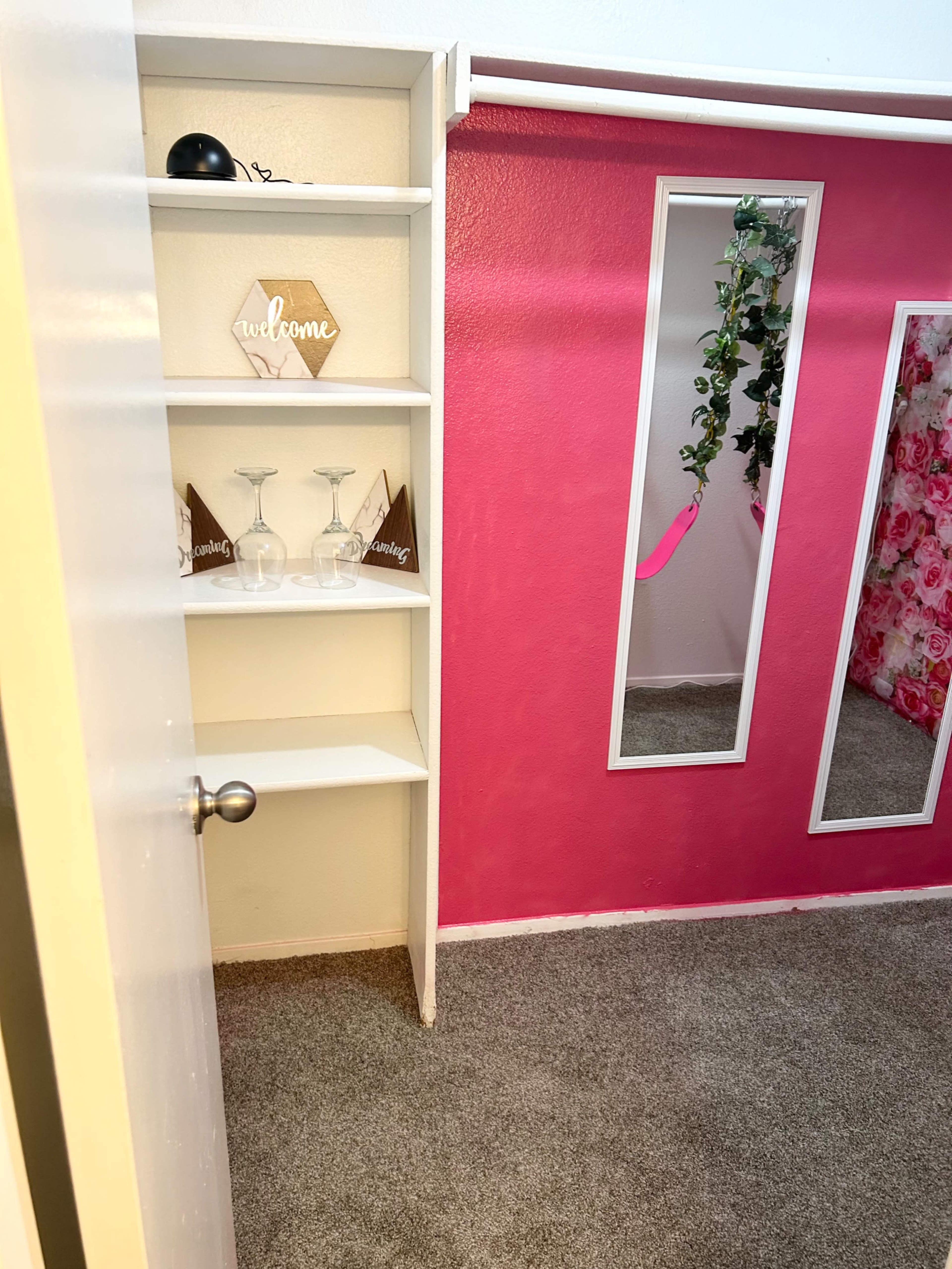 The image shows a corner of a room with a white shelving unit holding decorative items, a wall with a pink section and large mirrors, and a carpeted floor.