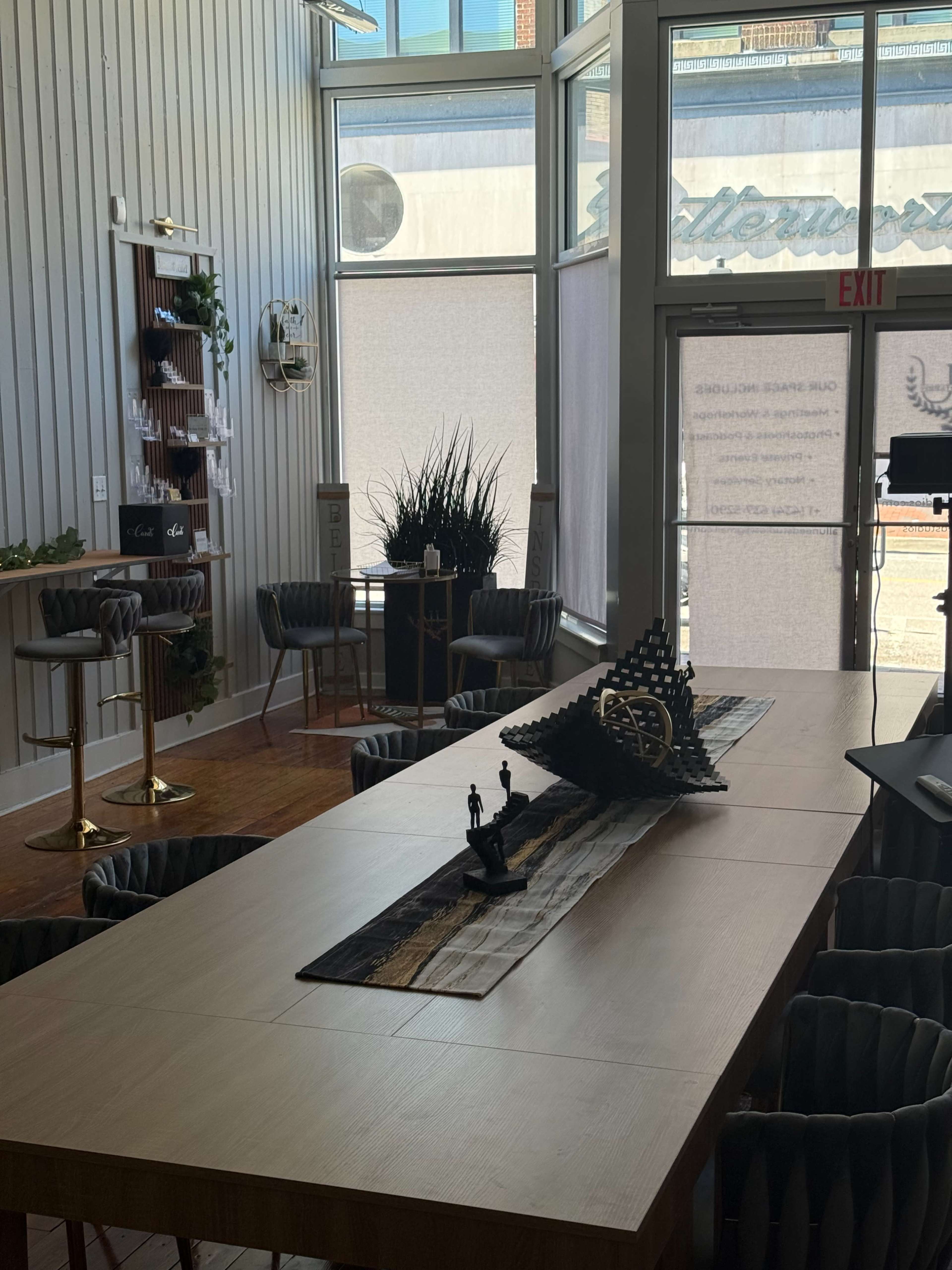 The image shows a modern interior space with a long wooden table, stylish chairs, and decorative elements like plants and sculptures, illuminated by natural light.