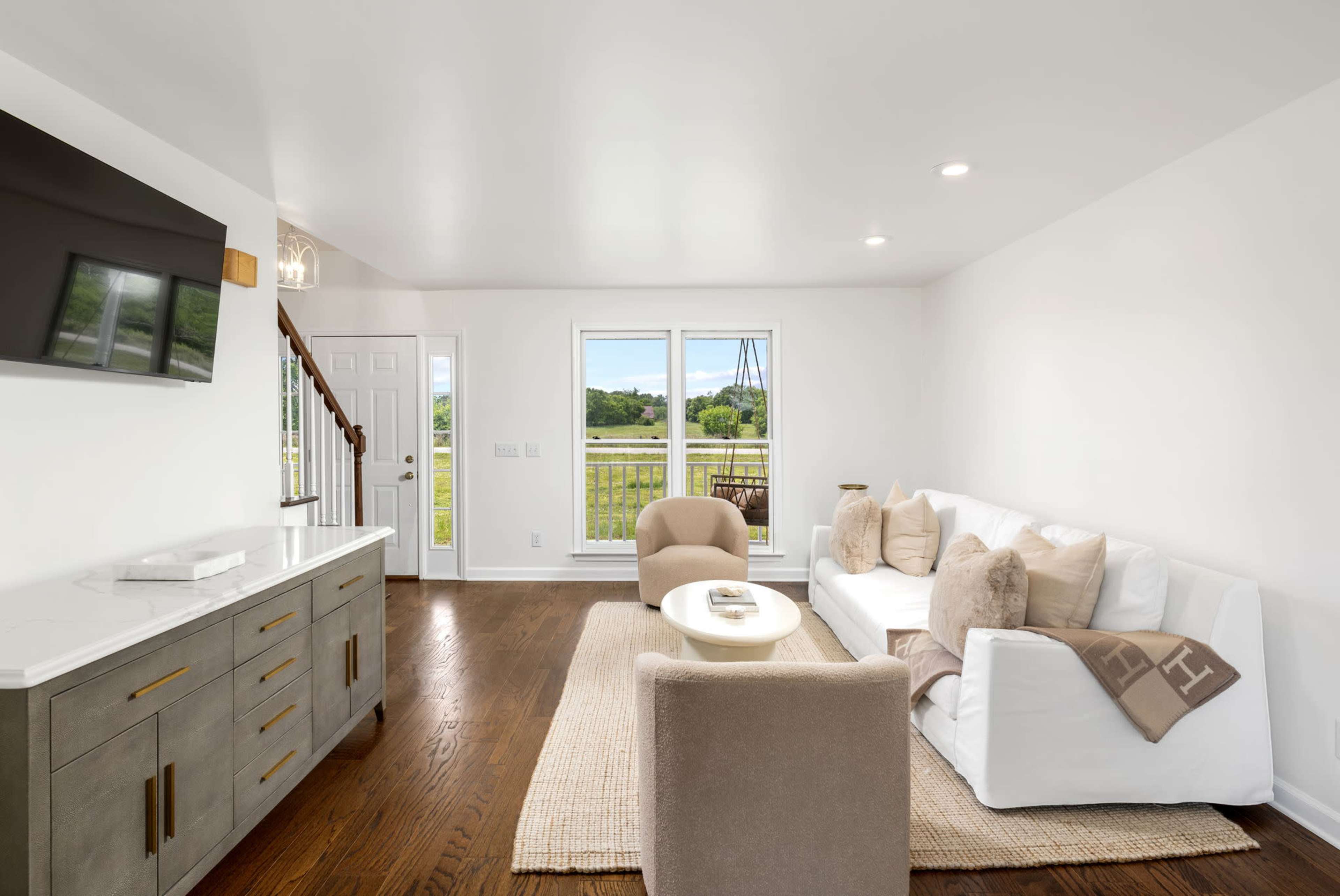 The image shows a bright living room featuring a white sofa, a beige armchair, a coffee table, and large windows that offer a view of the outdoors.