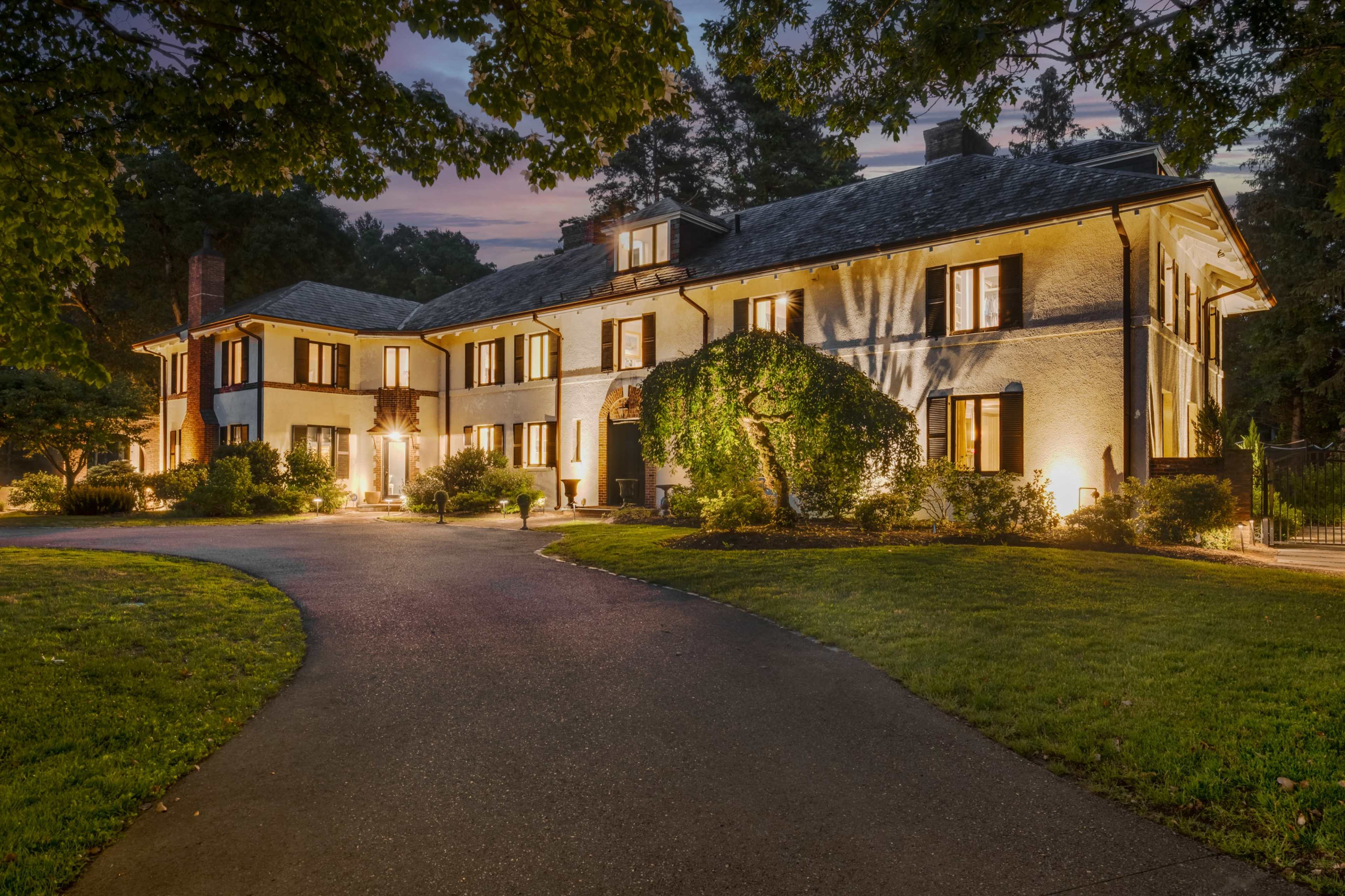 A large, two-story house with a landscaped front yard is illuminated at dusk, showcasing its windows and architectural details.