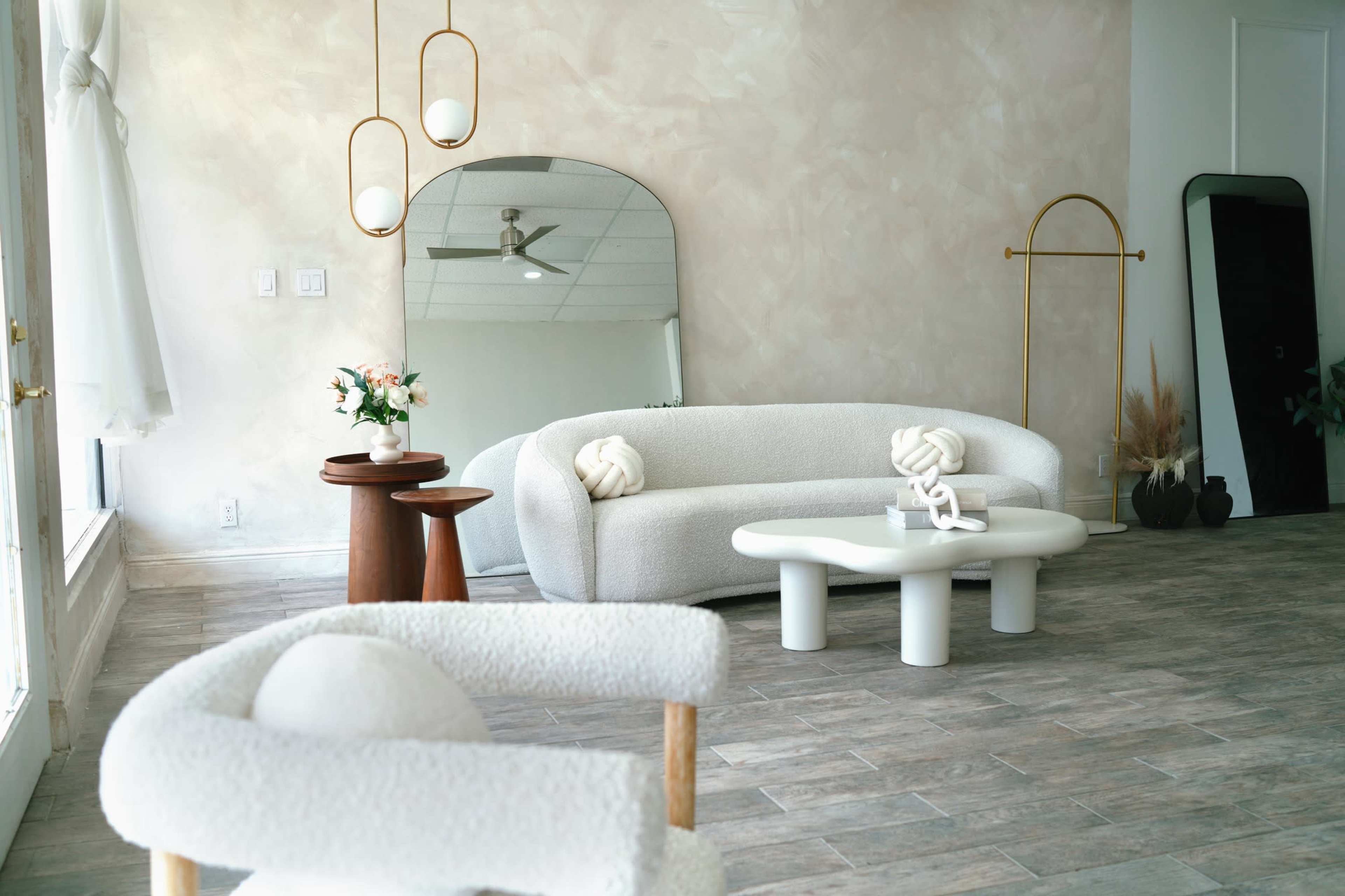 A modern interior space featuring a curved white sofa, a round wooden table, a low white coffee table, and decorative elements like flowers and mirrors against a softly colored wall.