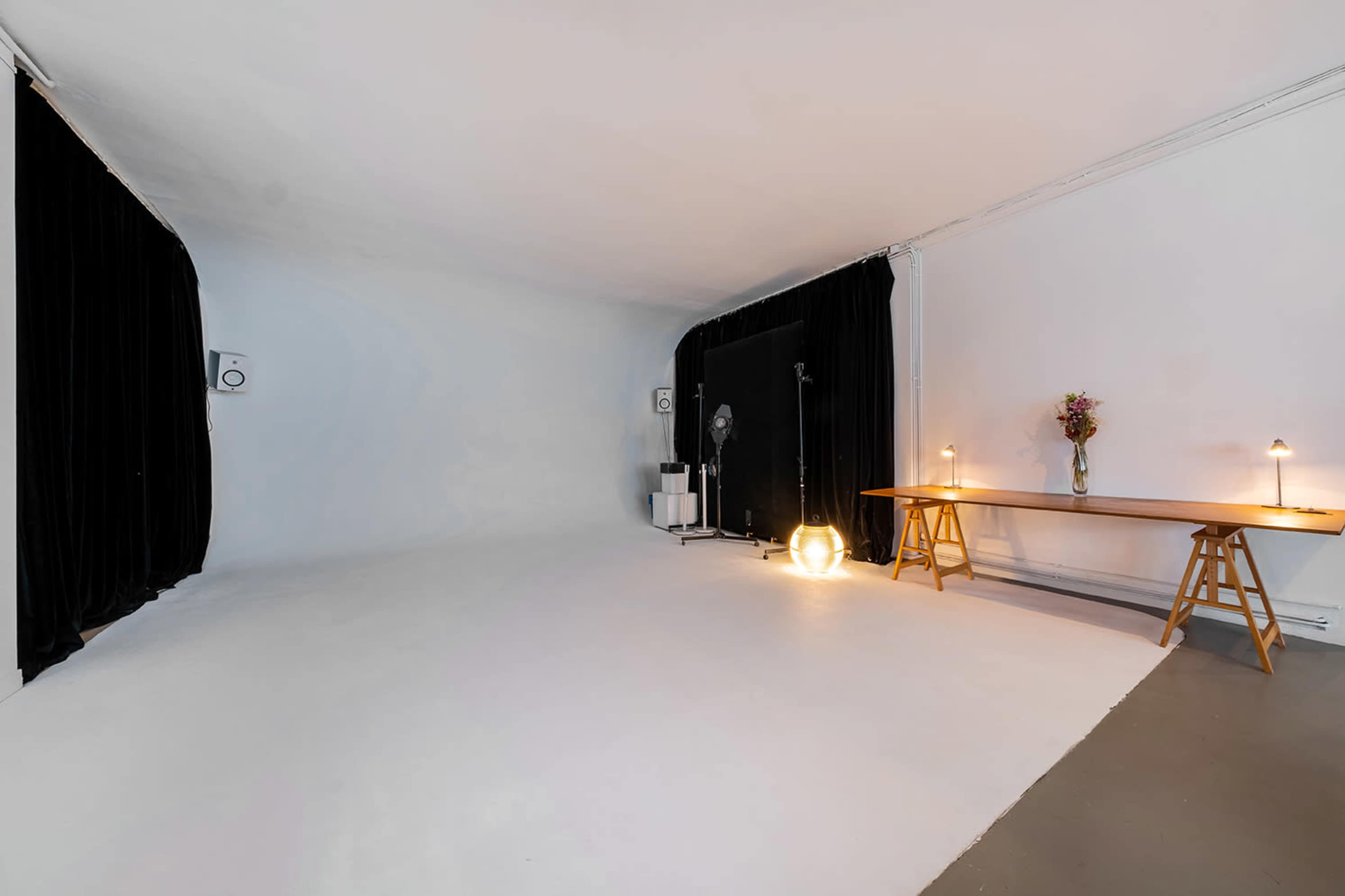A spacious, minimalist studio with a plain white backdrop, black curtains on one side, and a wooden table with lamps and a vase of flowers.