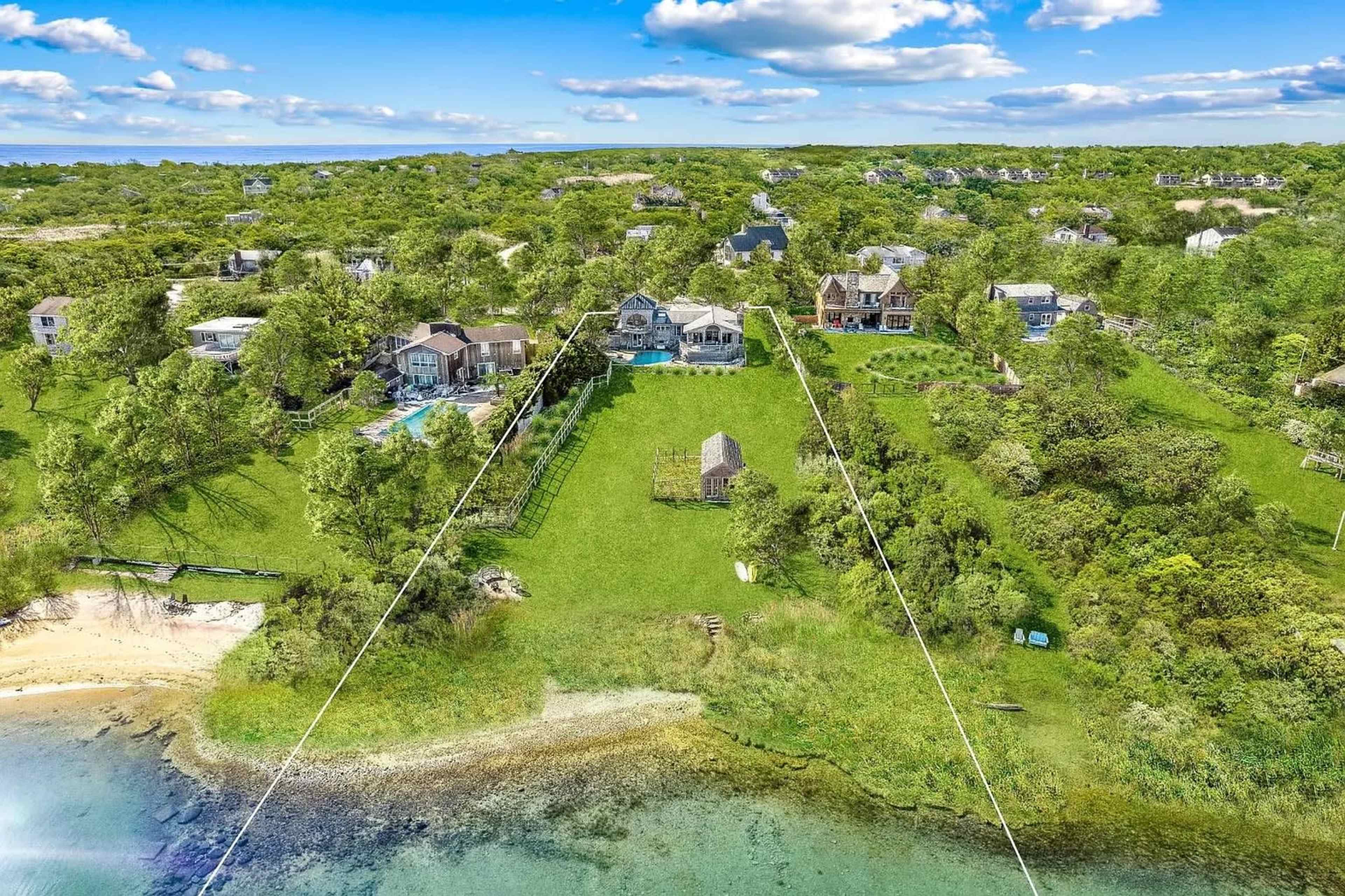 The image shows a coastal area with several houses, green lawns, and a waterfront, bordered by trees and blue skies.