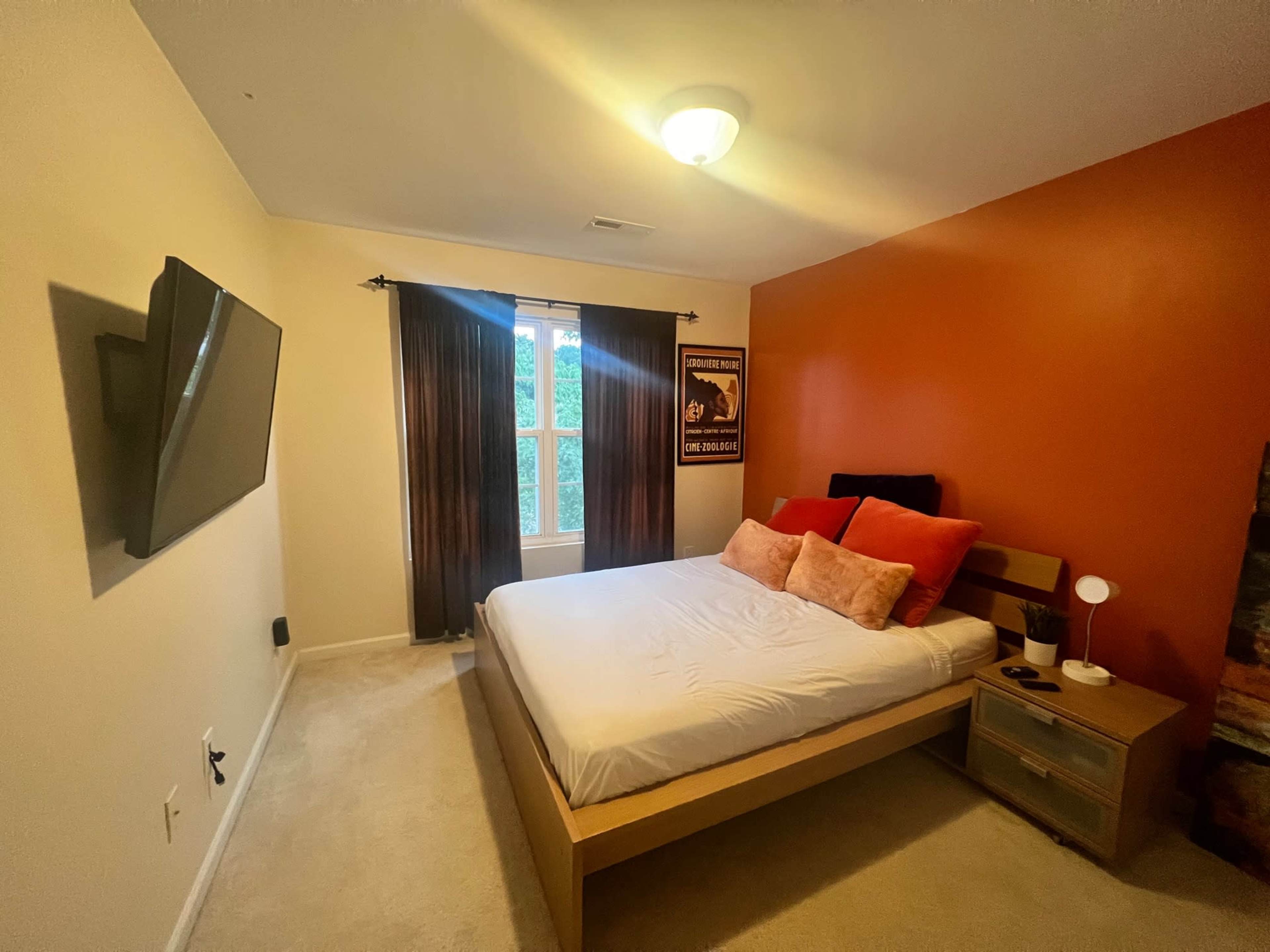 A bedroom features a double bed with orange accent wall, a window dressed with dark curtains, and a wall-mounted television.