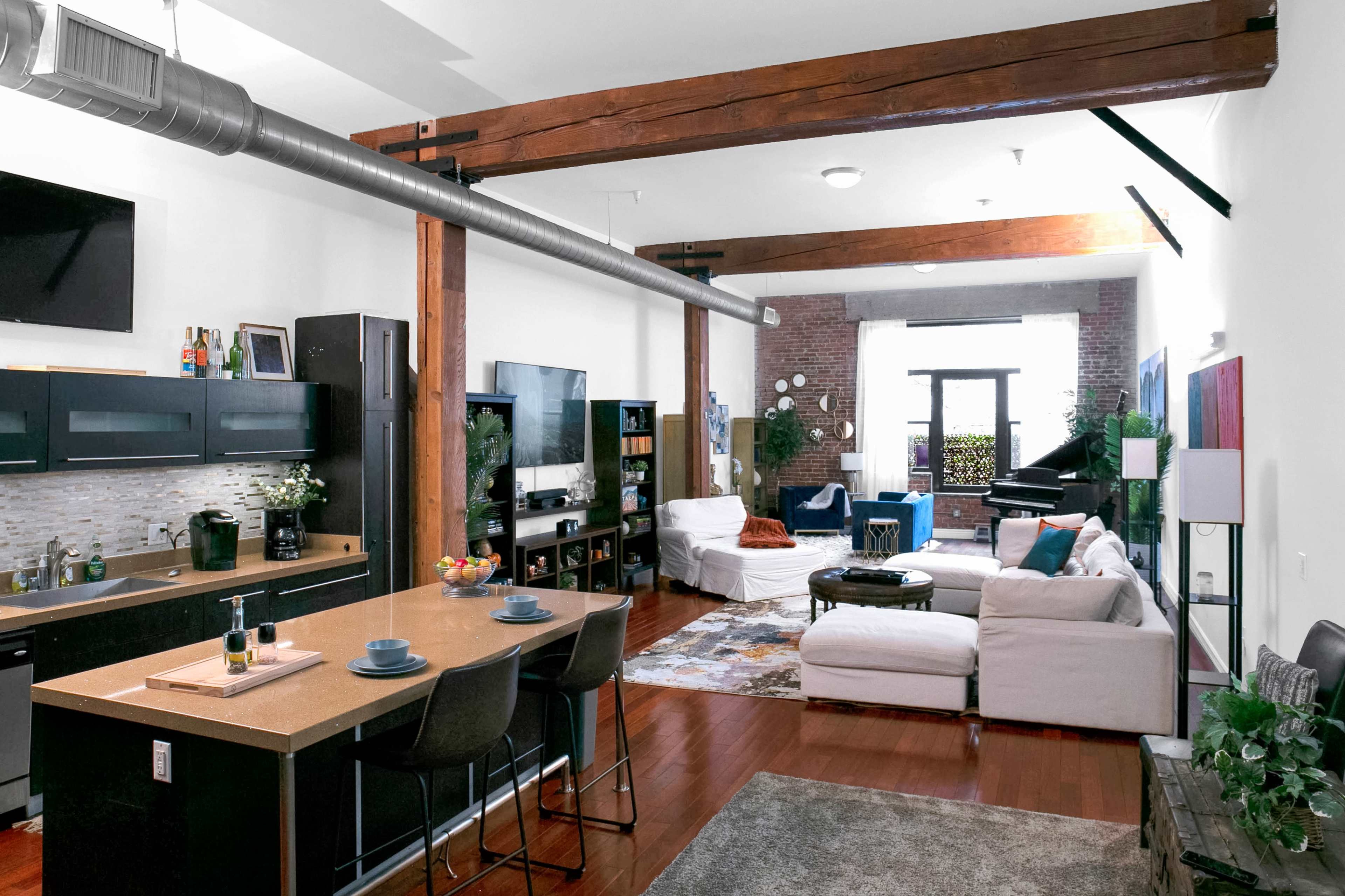 The image shows a modern loft-style living space featuring a kitchen with a breakfast bar, a cozy seating area, and large windows overlooking a balcony.