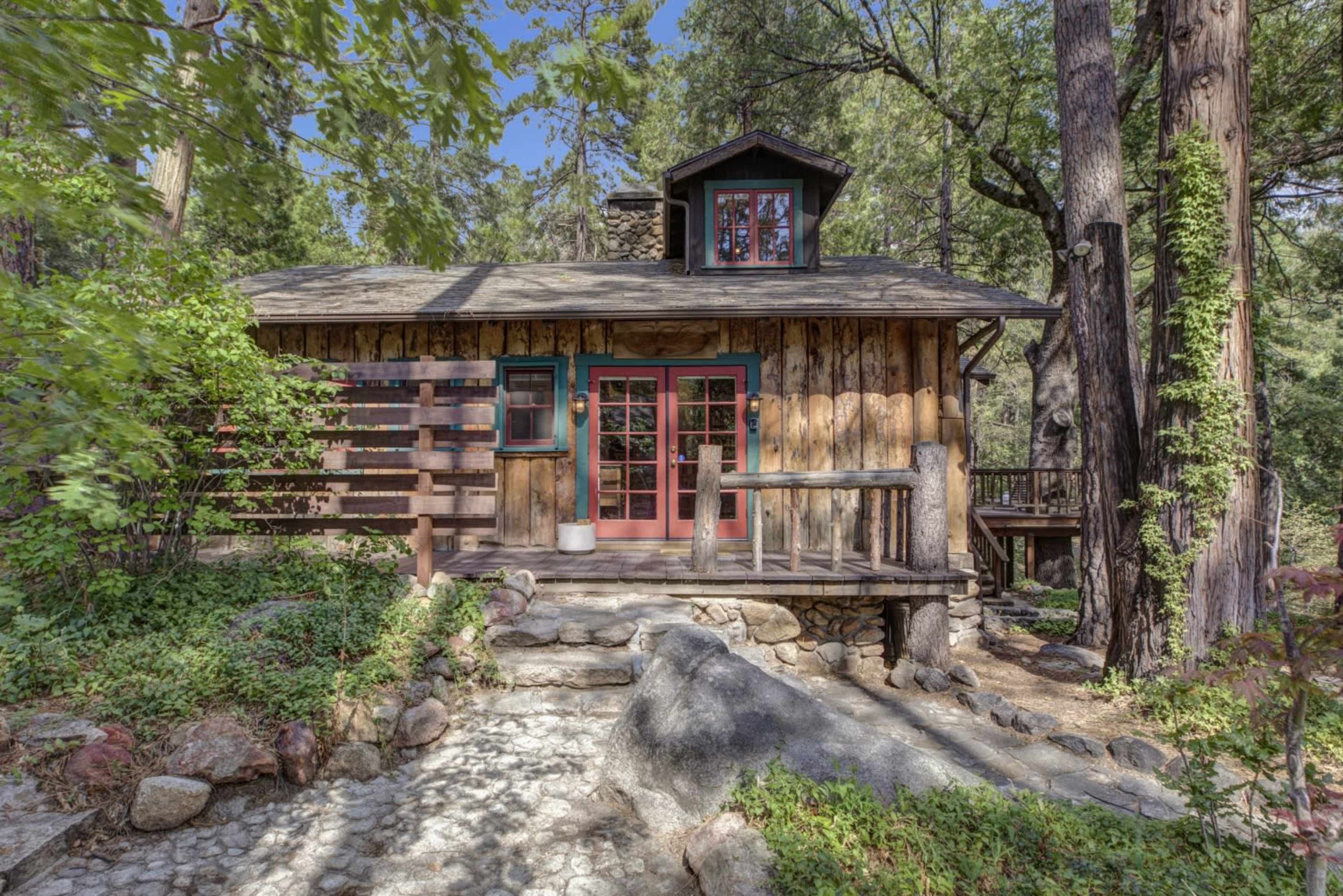 A rustic wooden cabin with a stone path leads to its front door, surrounded by tall trees and greenery.