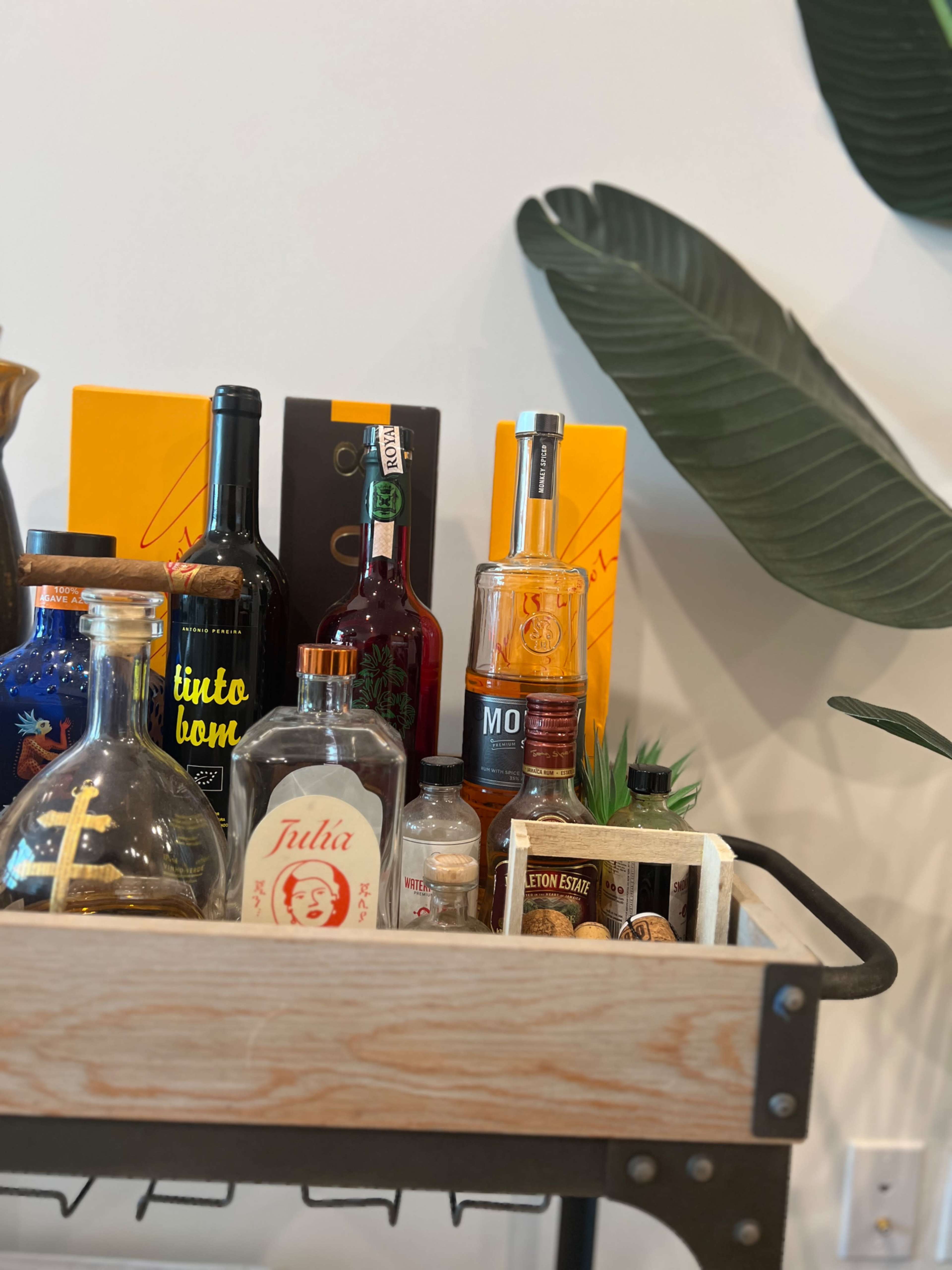 A bar cart is displayed with various bottles of liquor and decorative items against a wall with large green leaves in the background.