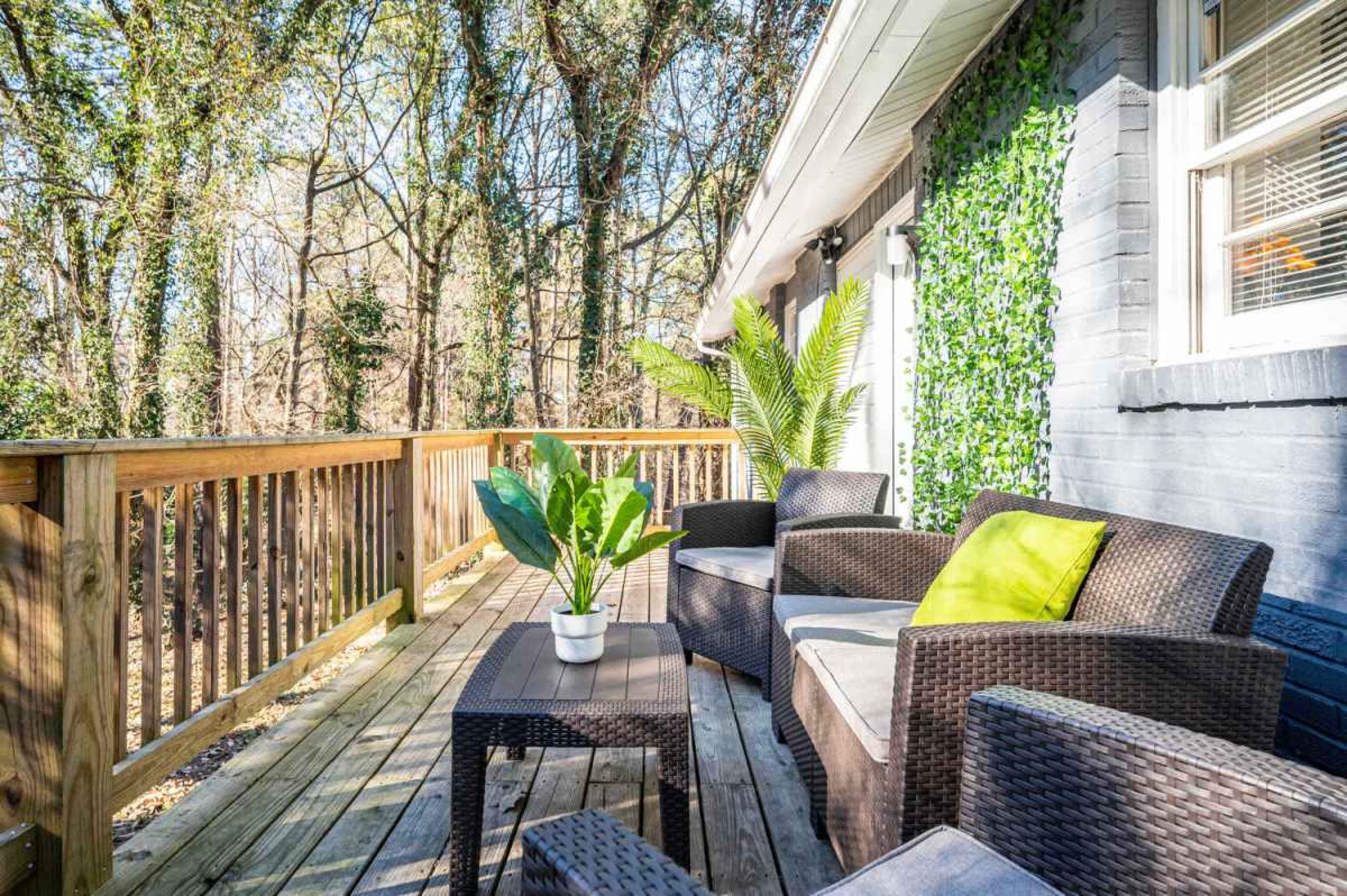 The image shows a wooden deck with wicker chairs and a table, surrounded by greenery and trees in the background.