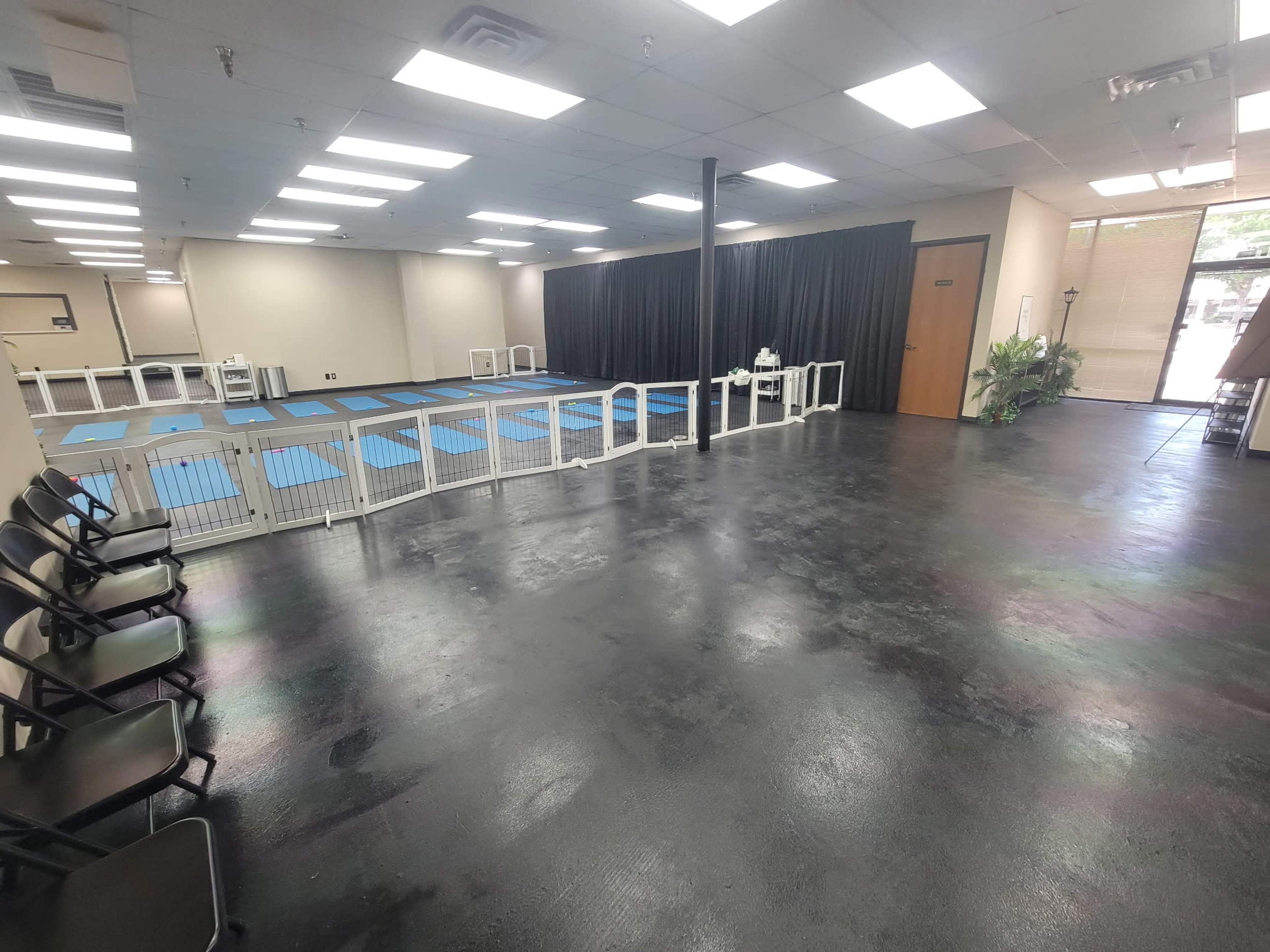 The image shows an empty training or activity space with blue mats, a seated area with chairs, and a black curtain at the far end.