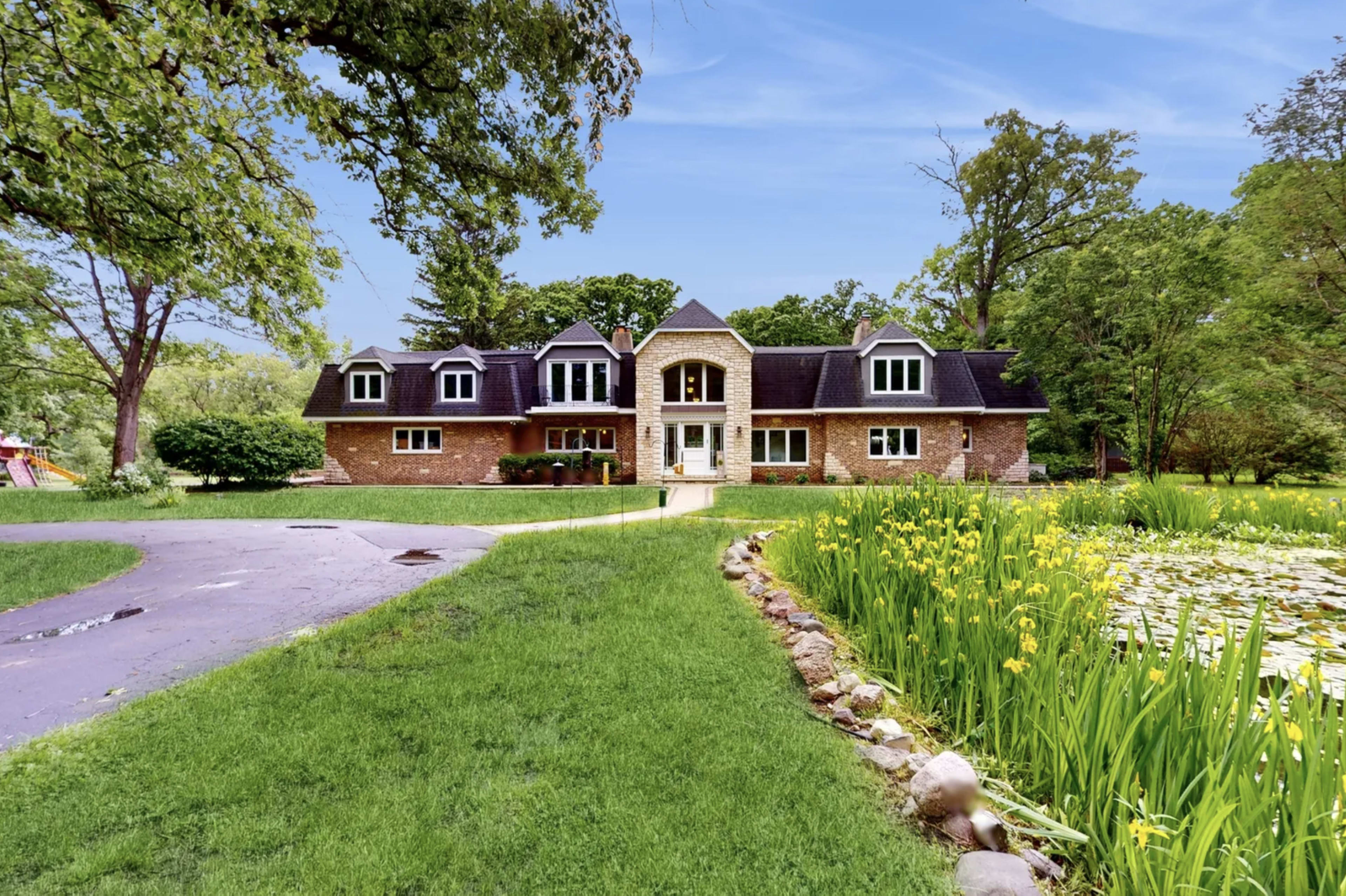 A large brick house with multiple gables sits beside a pond surrounded by greenery and flower patches.
